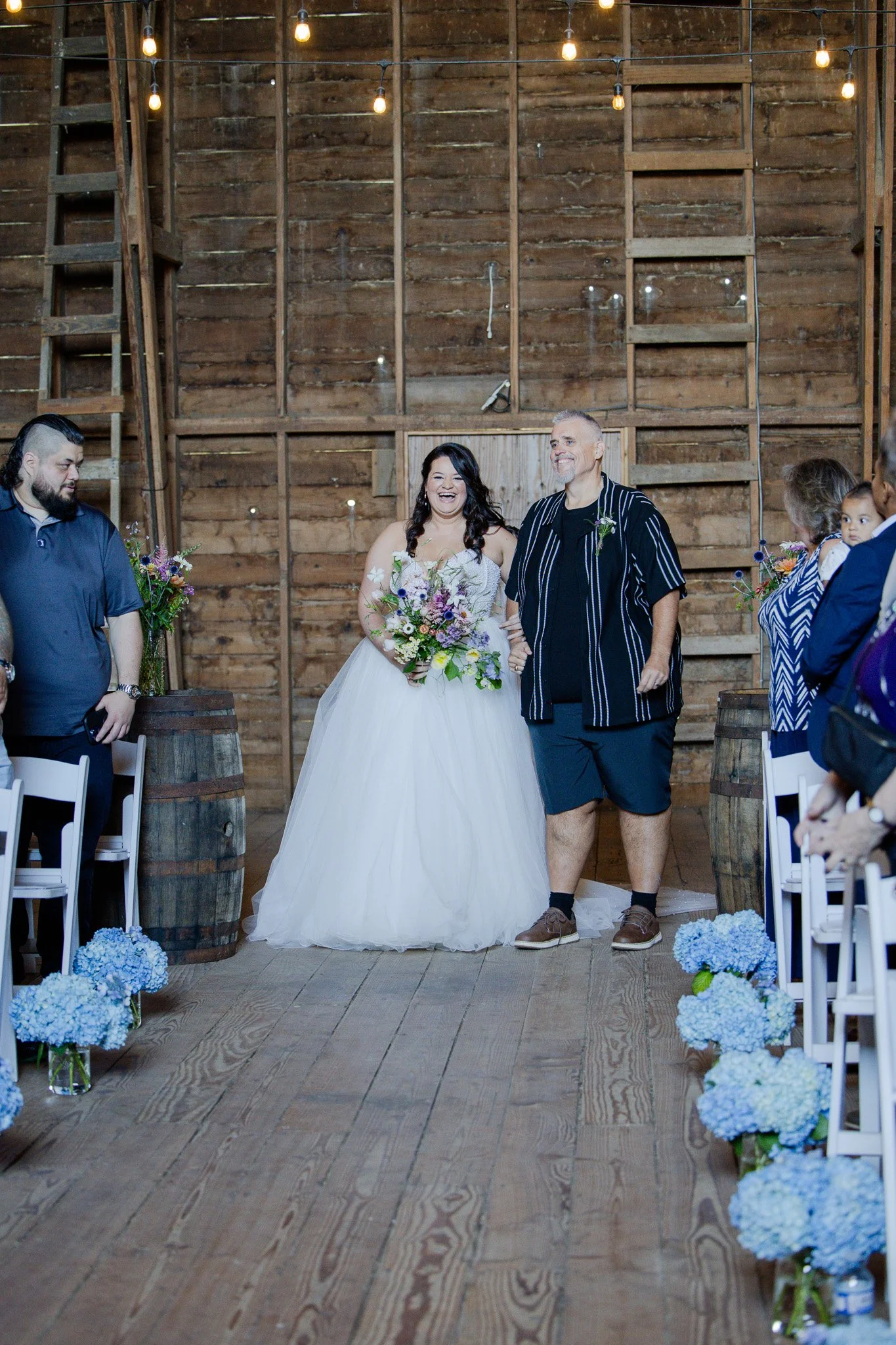 A bride in a white wedding dress holding a bouquet of flowers, walking arm-in-arm with a man in a black shirt and shorts, inside a rustic wooden venue with string lights and floral decorations.