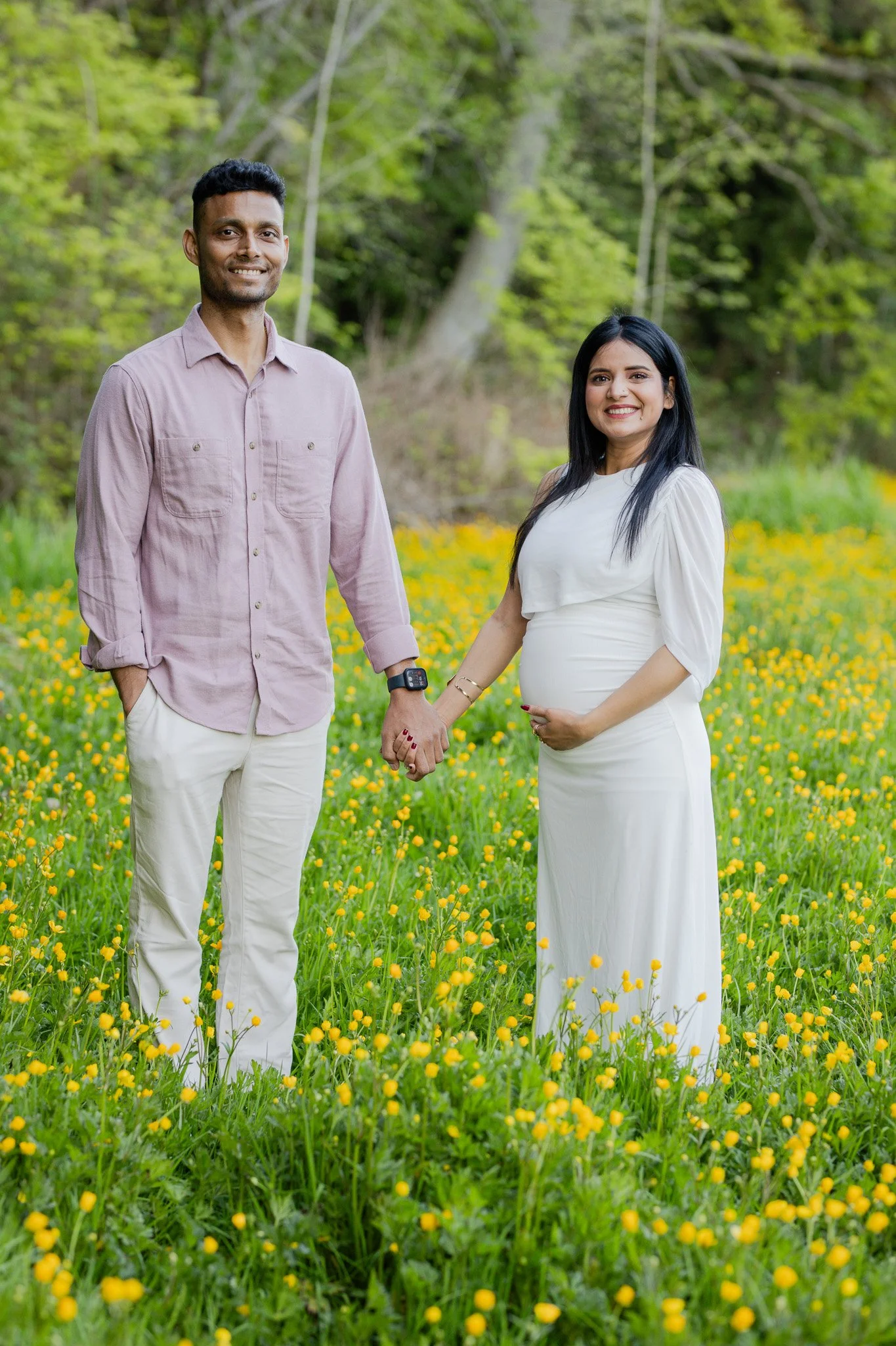 A smiling pregnant woman in a white dress holding hands with a smiling man in a light pink shirt and white pants, standing in a field of yellow flowers with green trees in the background.