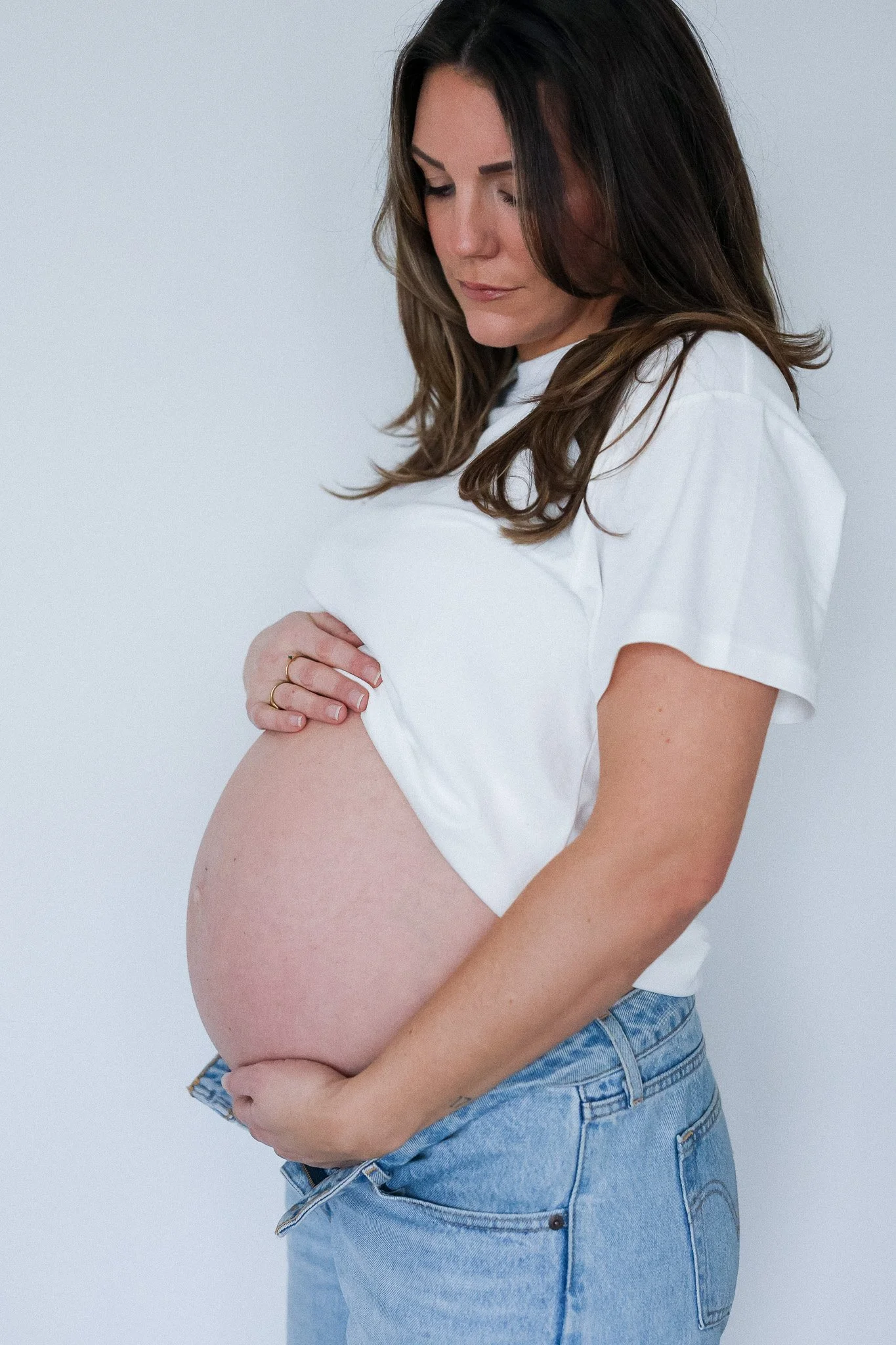 Pregnant woman with long brown hair wearing a white T-shirt and blue jeans, looking down at her pregnant belly in a bright studio with a plain white background.