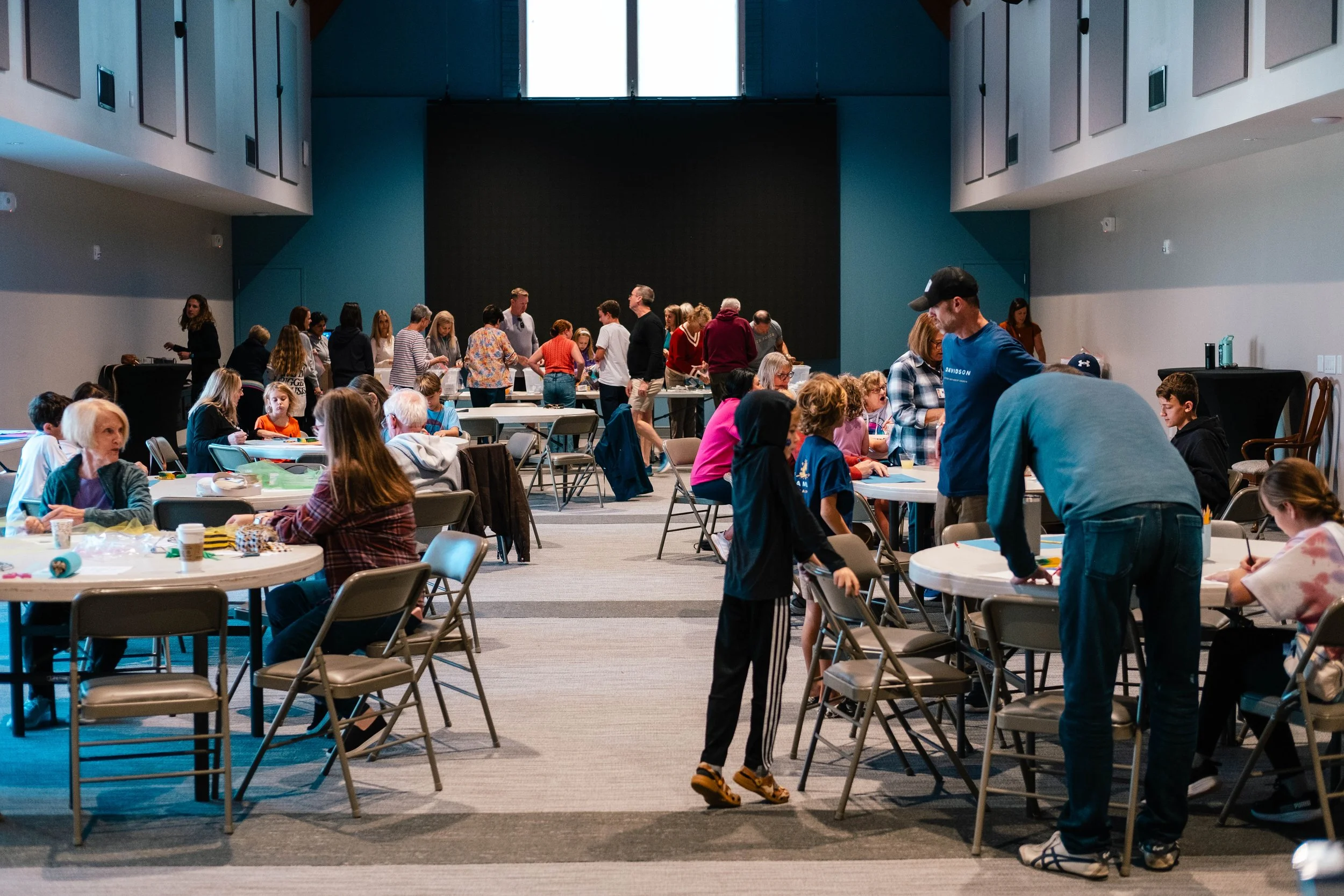 People gathered in a large conference room participating in a group activity, with some seated at tables and others standing or walking around.
