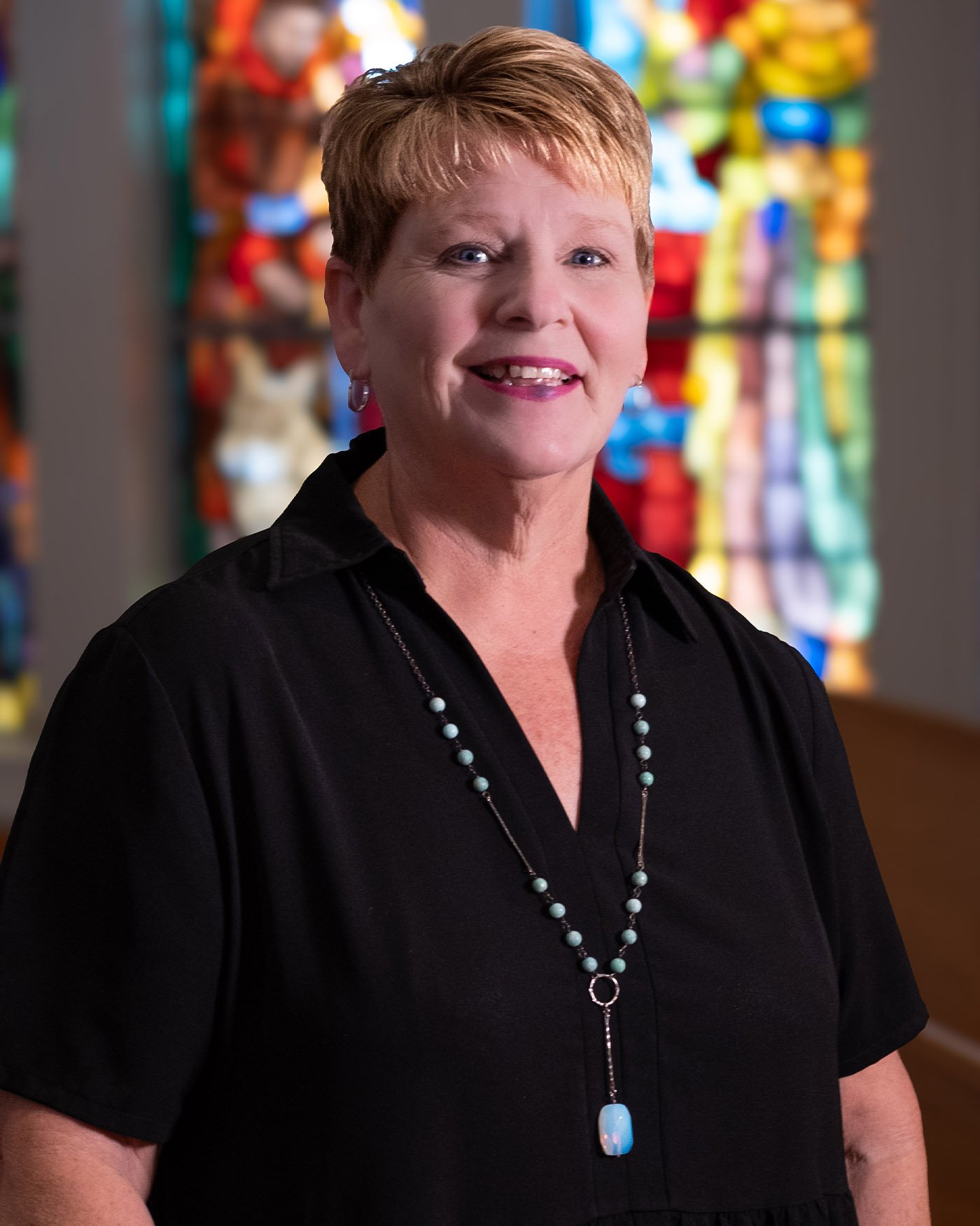 A smiling woman with short reddish hair, wearing a black blouse and beaded necklace, standing in front of colorful yarns or thread displays.