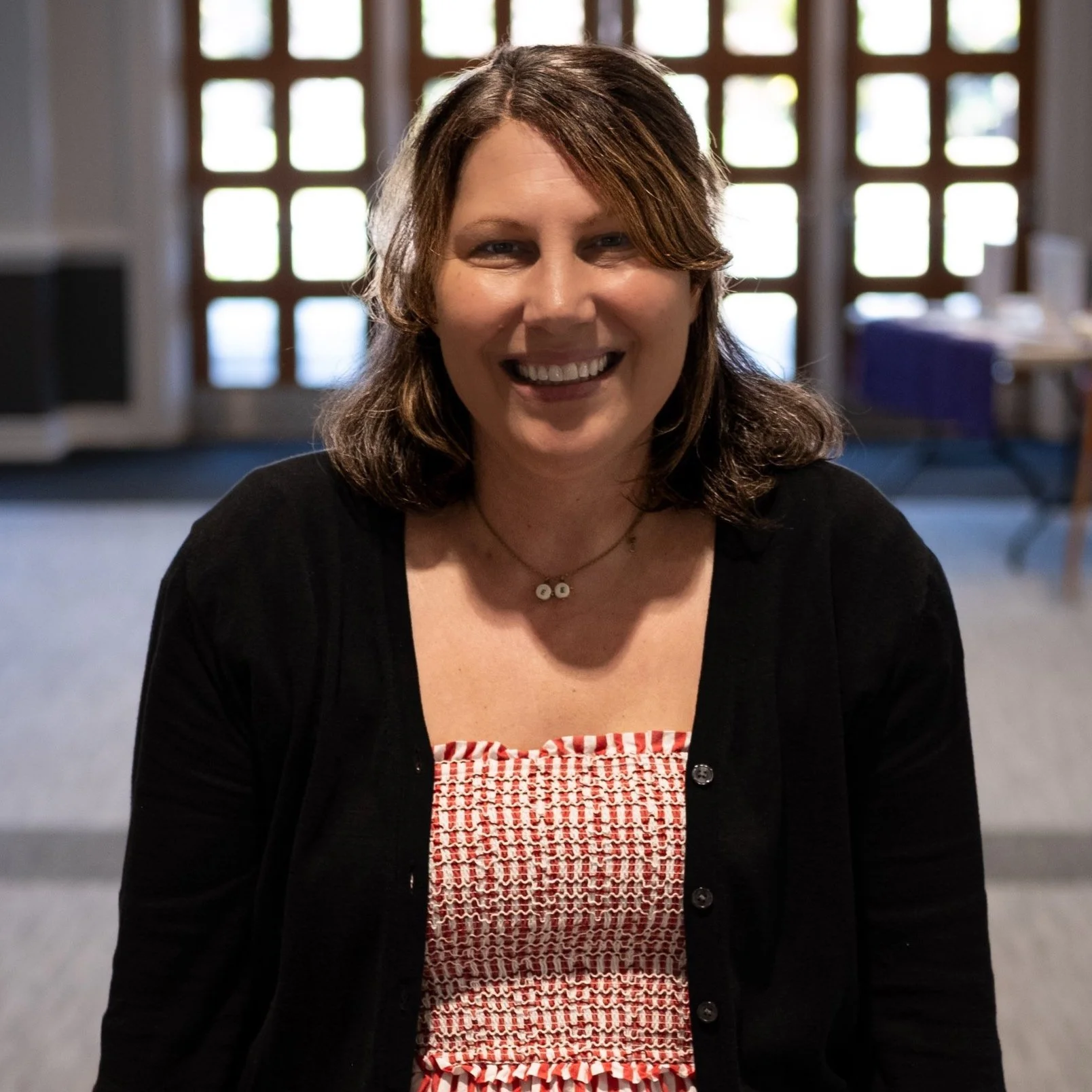 A woman with shoulder-length brown hair smiling at the camera, wearing a black cardigan and a red and white patterned top, inside a room with a large window with grid-like panes in the background.
