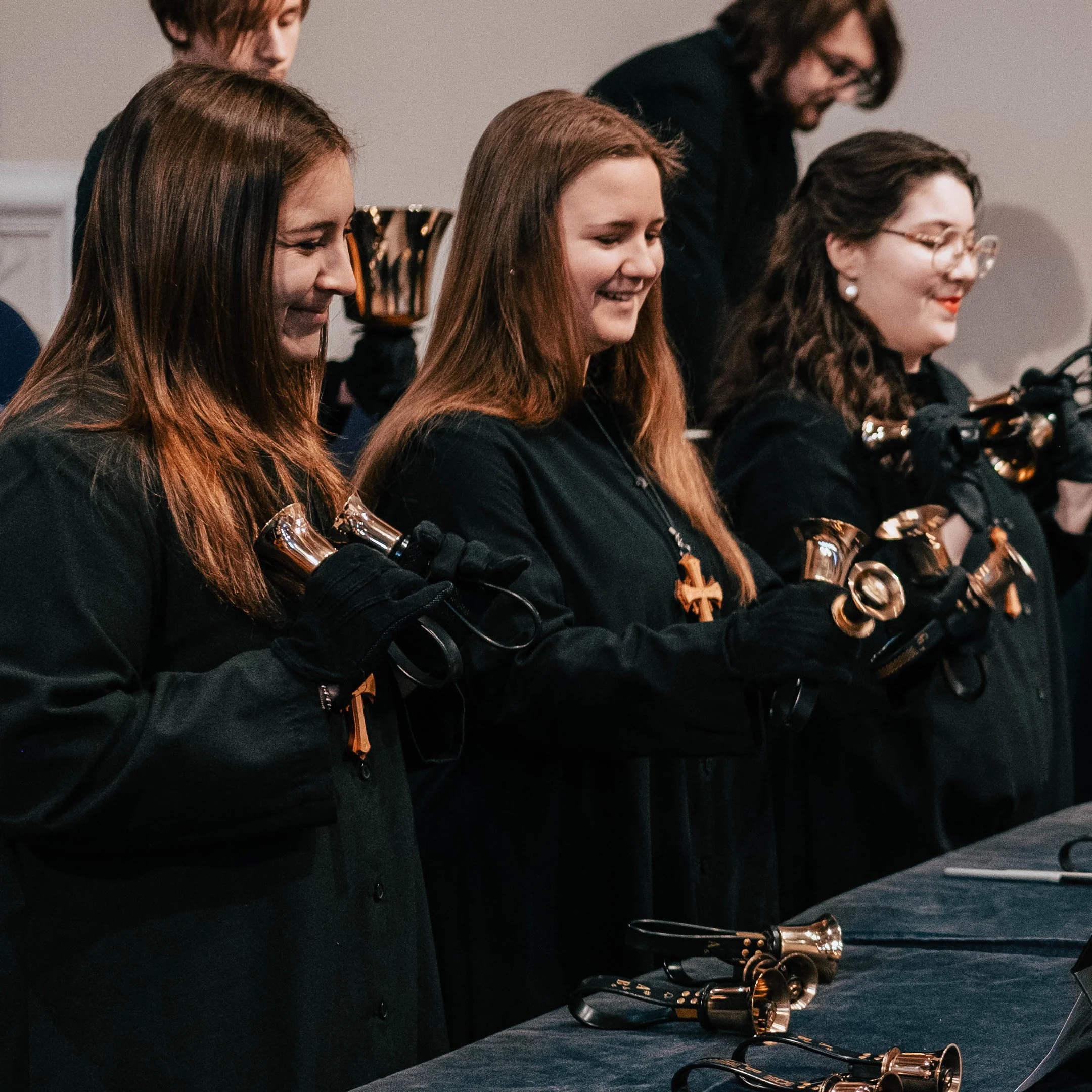 Four women in black robes and gloves holding bells, participating in a religious ceremony, with others in the background.