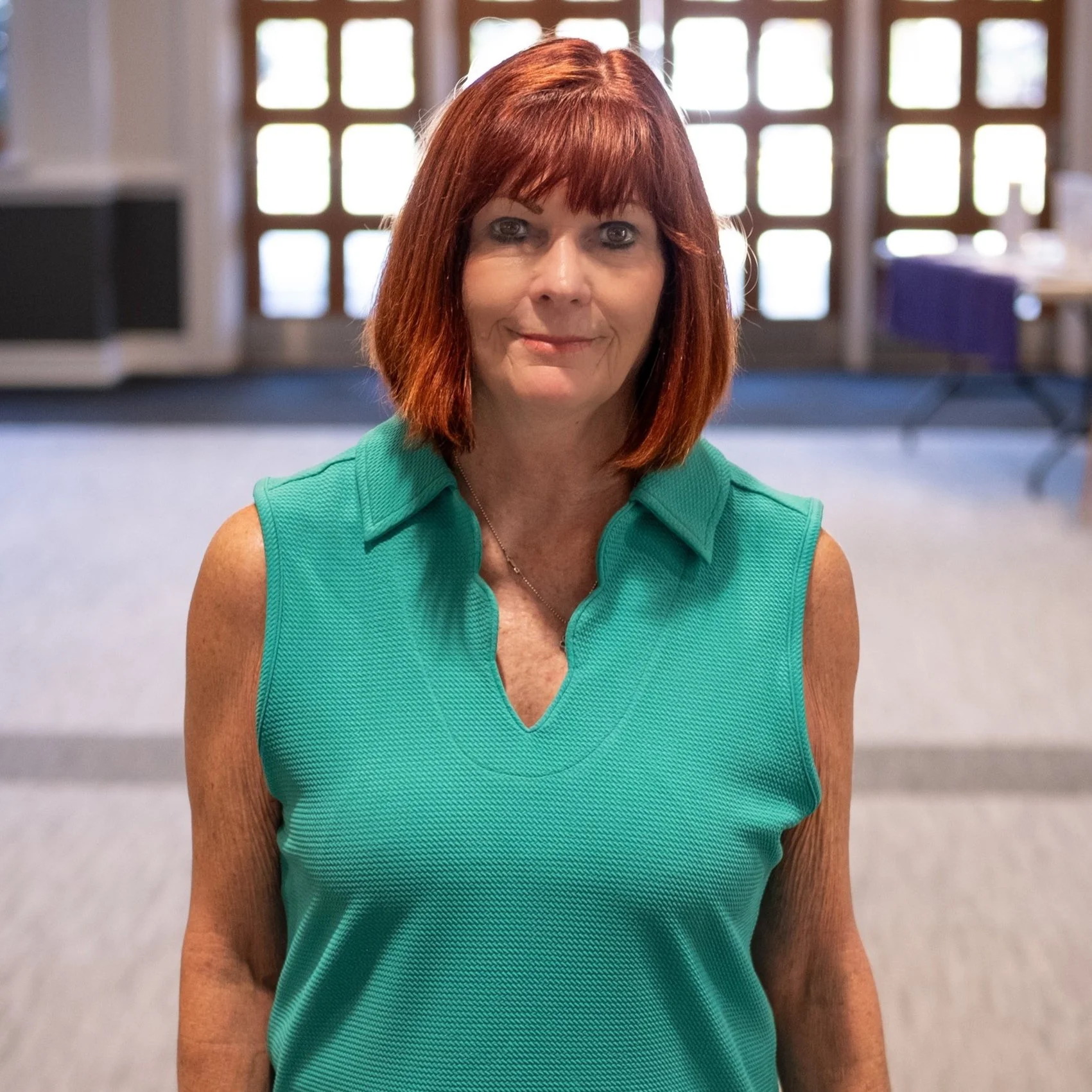 A woman with red hair wearing a sleeveless teal top standing indoors with a large window in the background.
