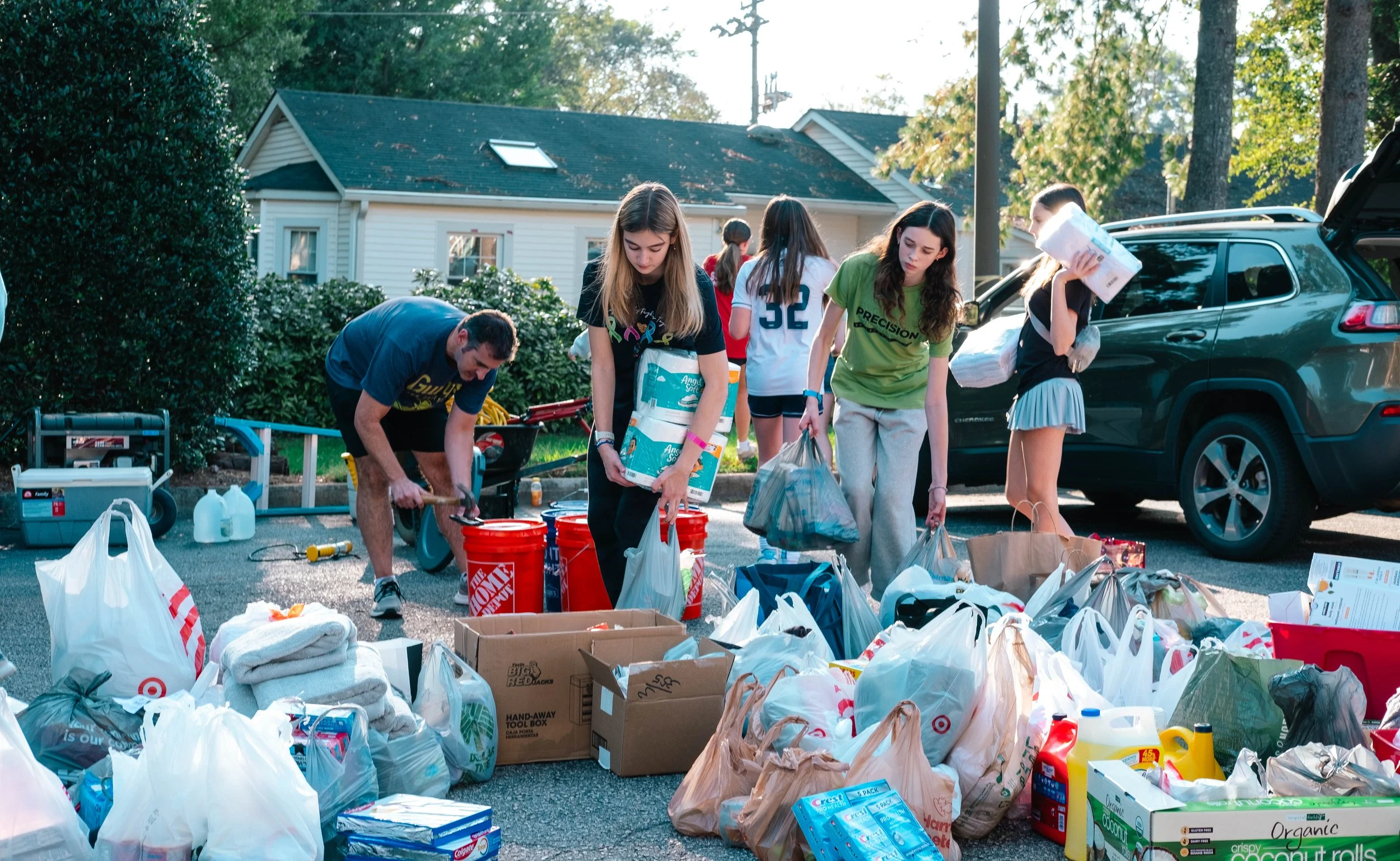 People sorting and organizing donations on a driveway, with bags and boxes of goods, some with household items like towels and cleaning supplies, and a person carrying a large pack of paper towels.