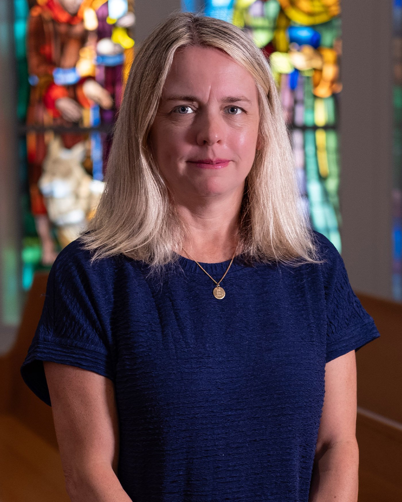 A woman with blonde hair wearing a navy blue top and a gold necklace, standing indoors with colorful stained glass in the background.