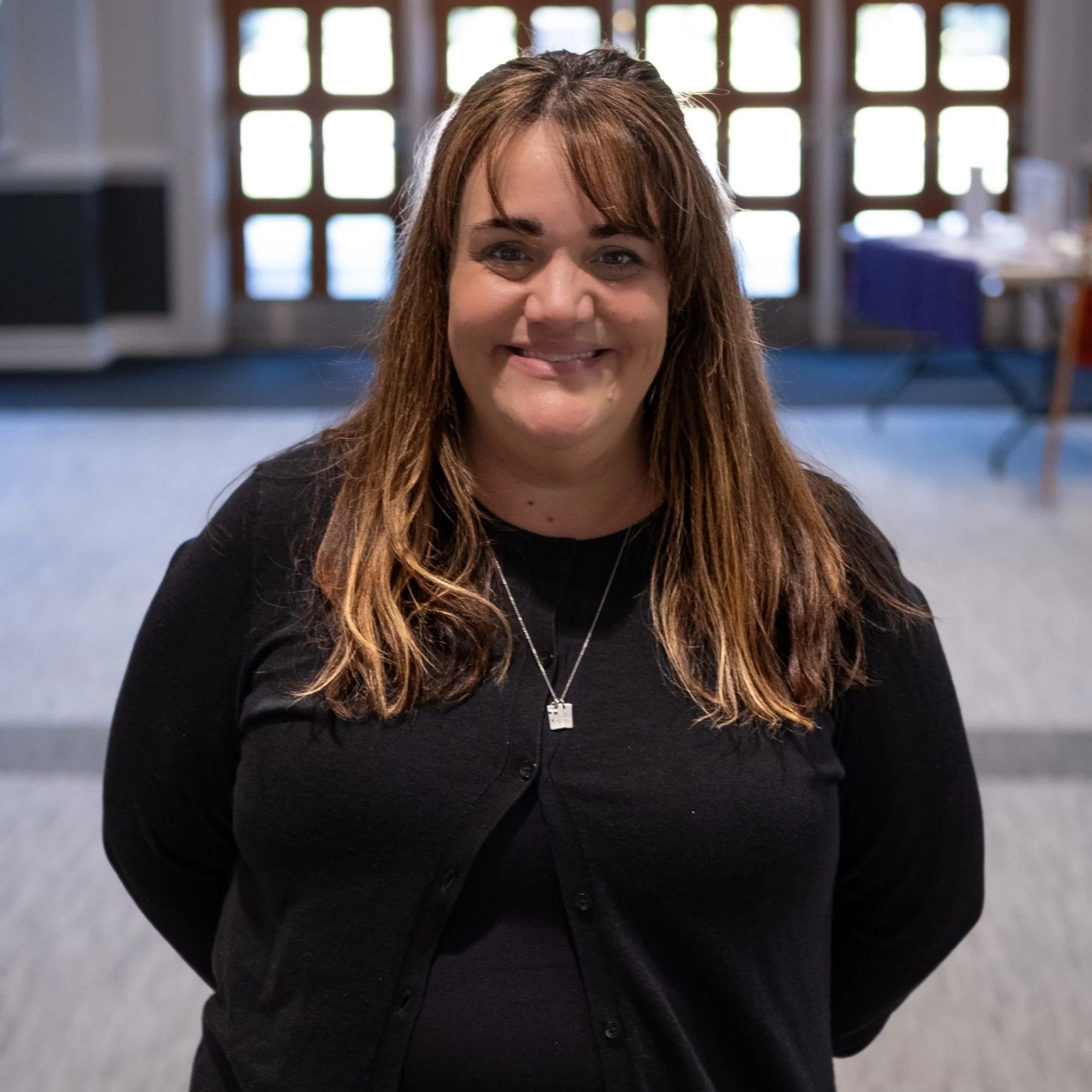 A woman with long brown hair and a black cardigan smiling in an indoor setting with windows and furniture in the background.