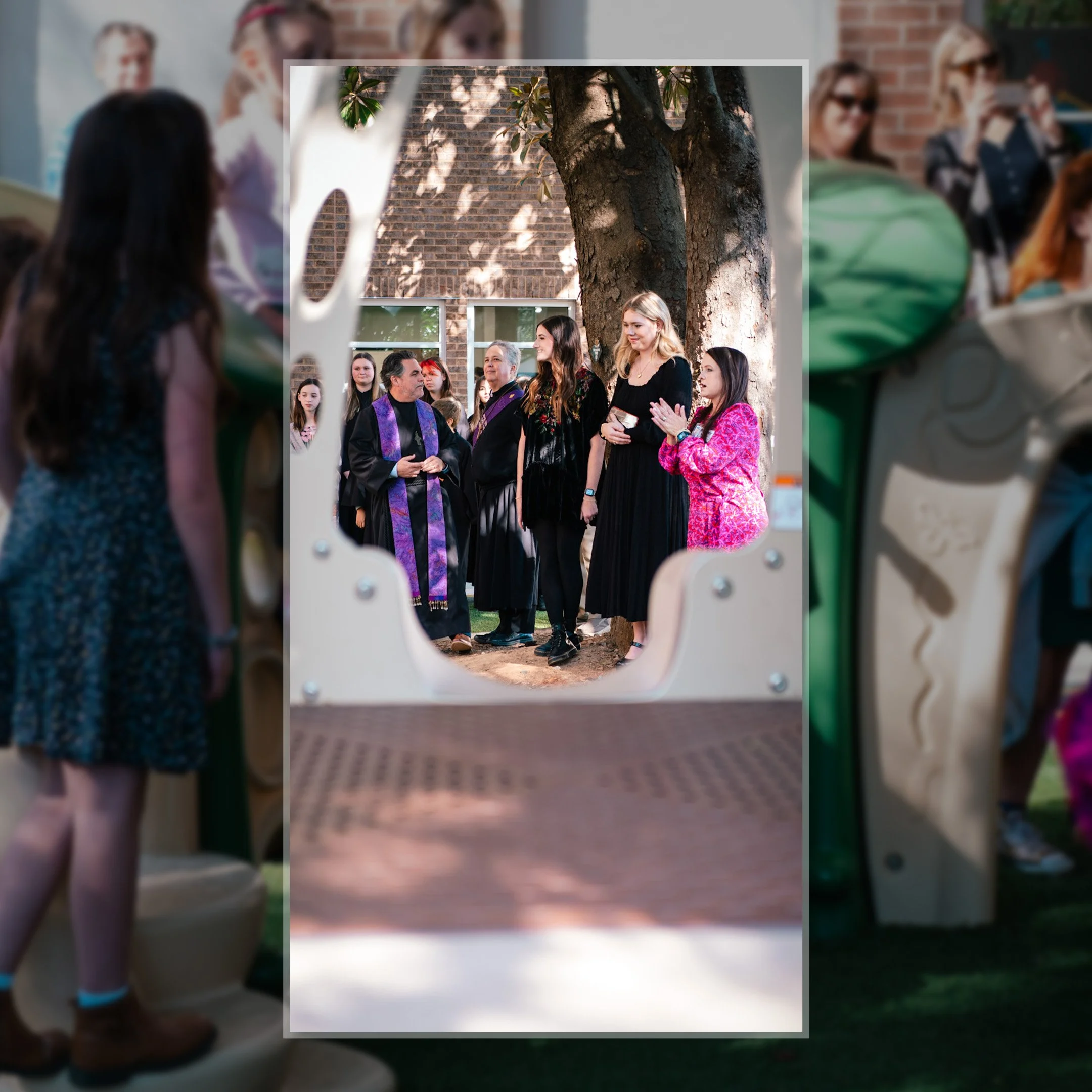 People participating in a ceremony outdoors, some dressed in formal black, with one person wearing a purple clergy stole, gathered around a tree, with others clapping and watching.