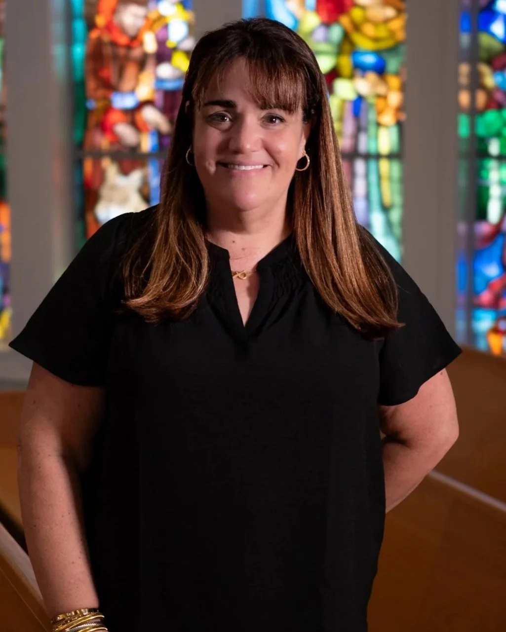 A woman with long brown hair and a black cardigan smiling in an indoor setting with windows and furniture in the background.