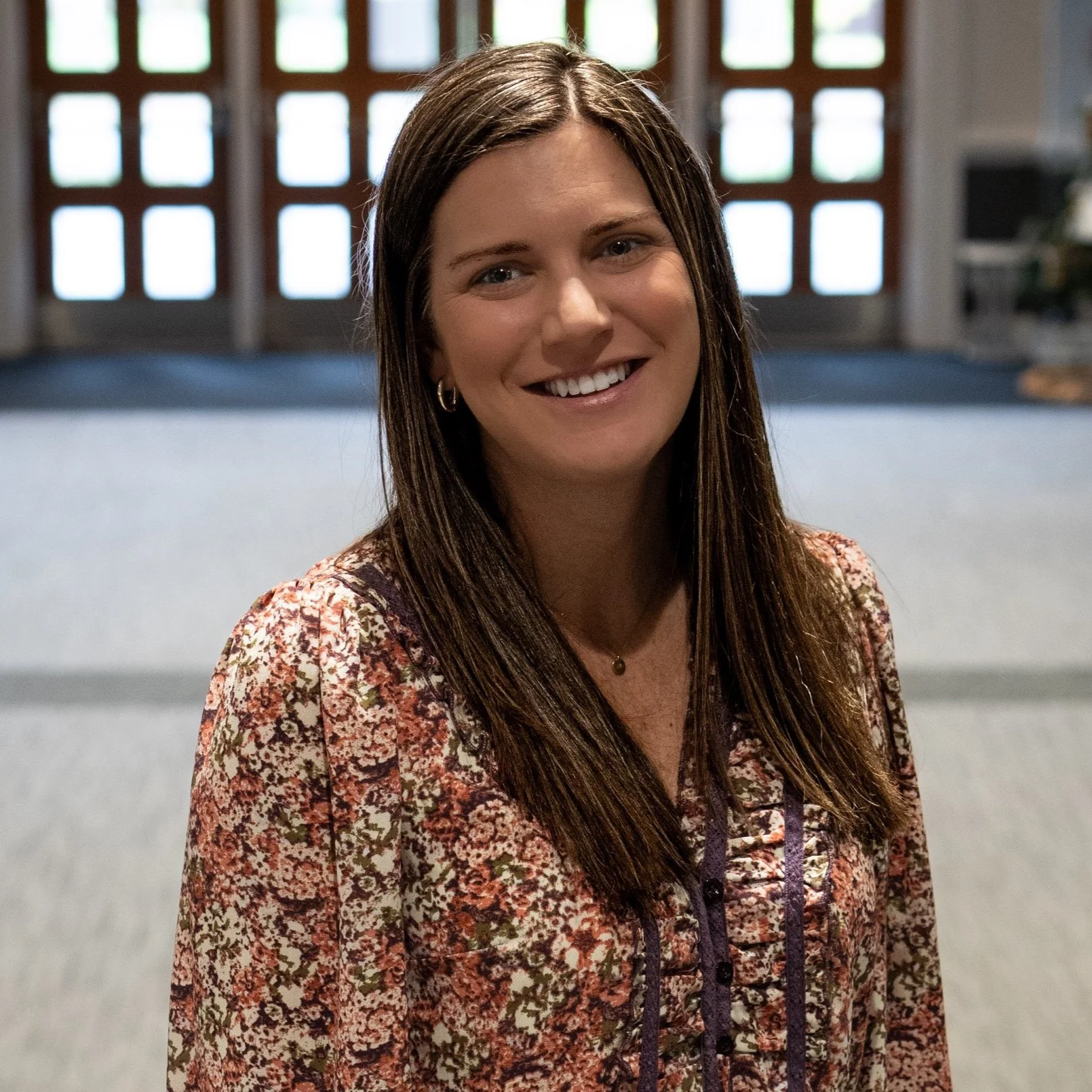 A woman with long brown hair smiling indoors, standing in front of a window with a geometric pattern.