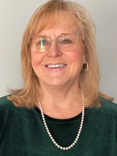 A smiling middle-aged woman with shoulder-length blonde hair, wearing glasses, earrings, a pearl necklace, and a dark green top, standing against a plain light-colored wall.