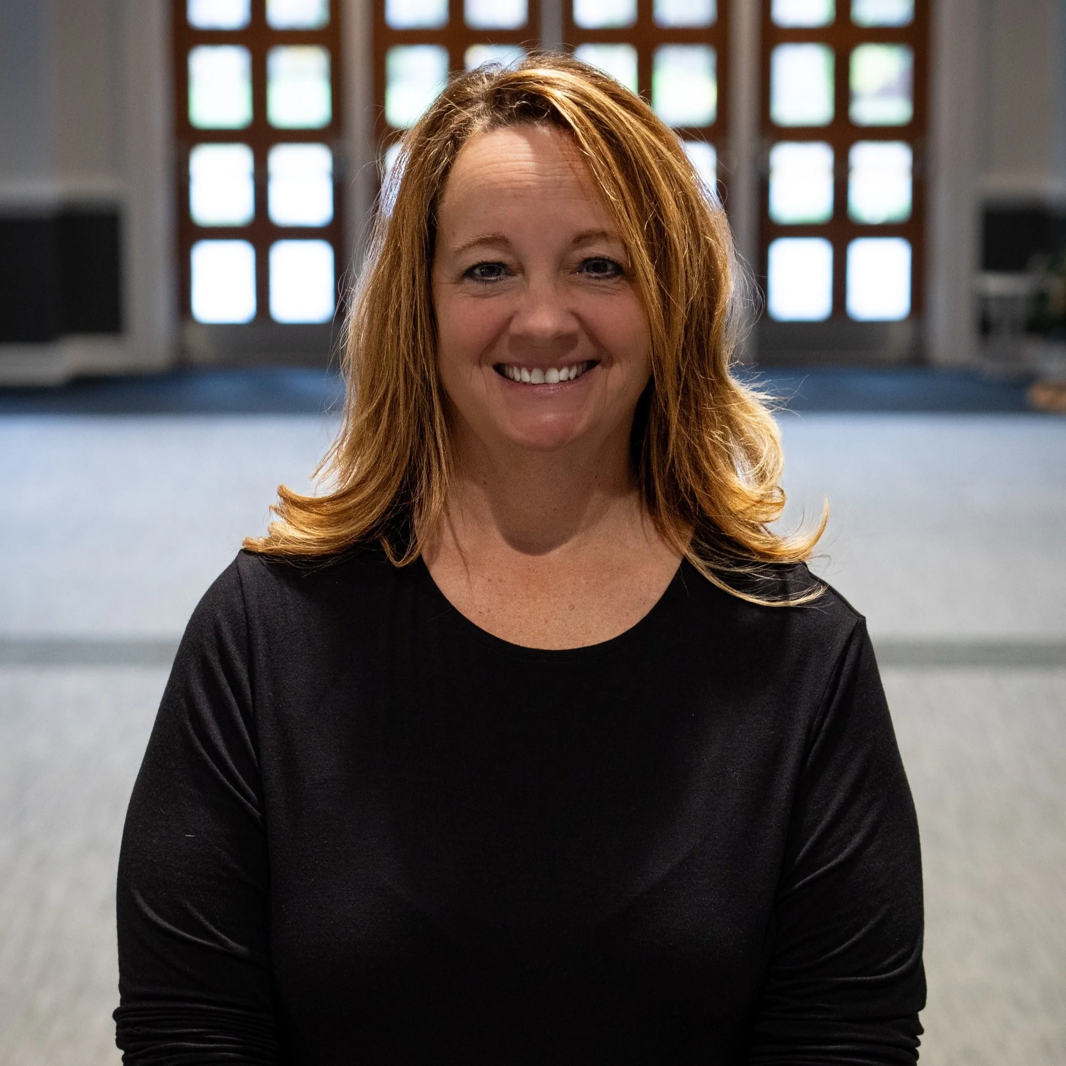 A woman with red hair smiling, wearing a black shirt, in an indoor setting with large windows in the background.