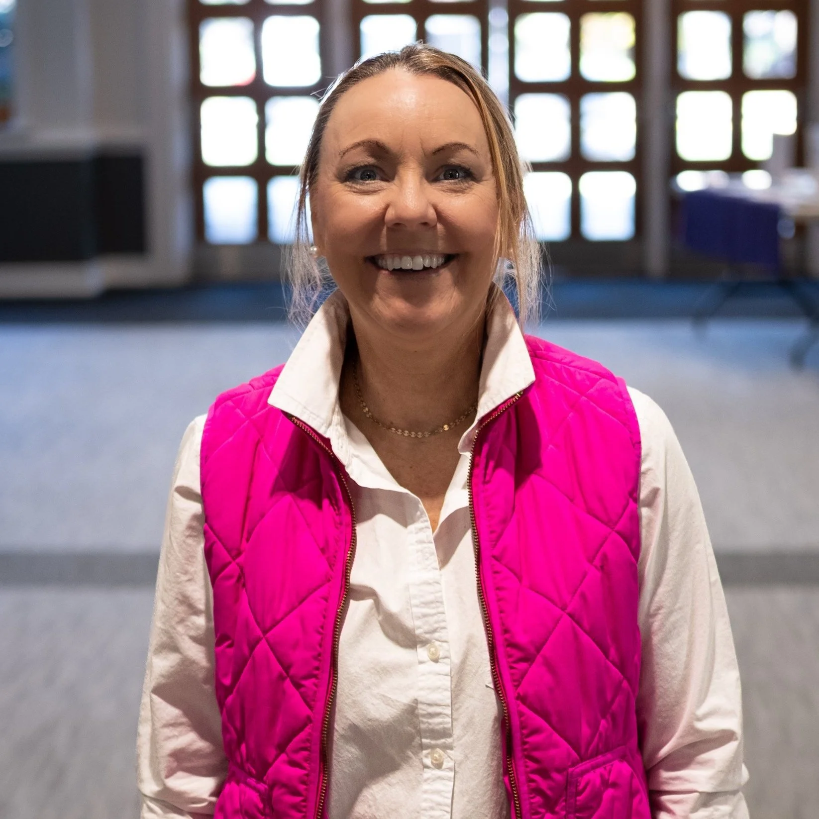 Smiling woman with light brown hair in a ponytail, wearing a white button-up shirt and a pink quilted vest, standing inside a building with large window panes in the background.