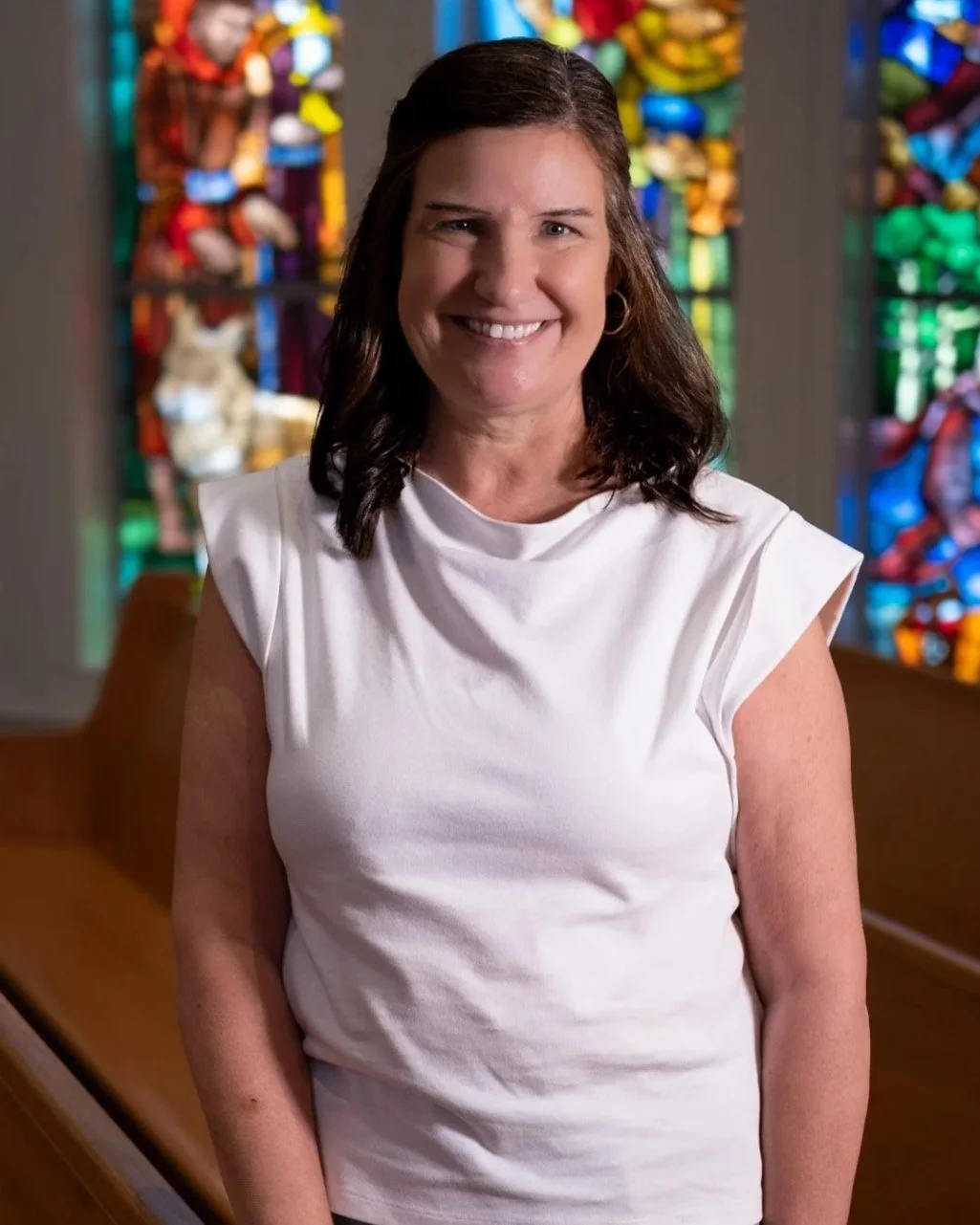 A smiling woman with shoulder-length dark hair, wearing a magenta blouse, standing indoors in front of large windows with lattice-patterned frames.