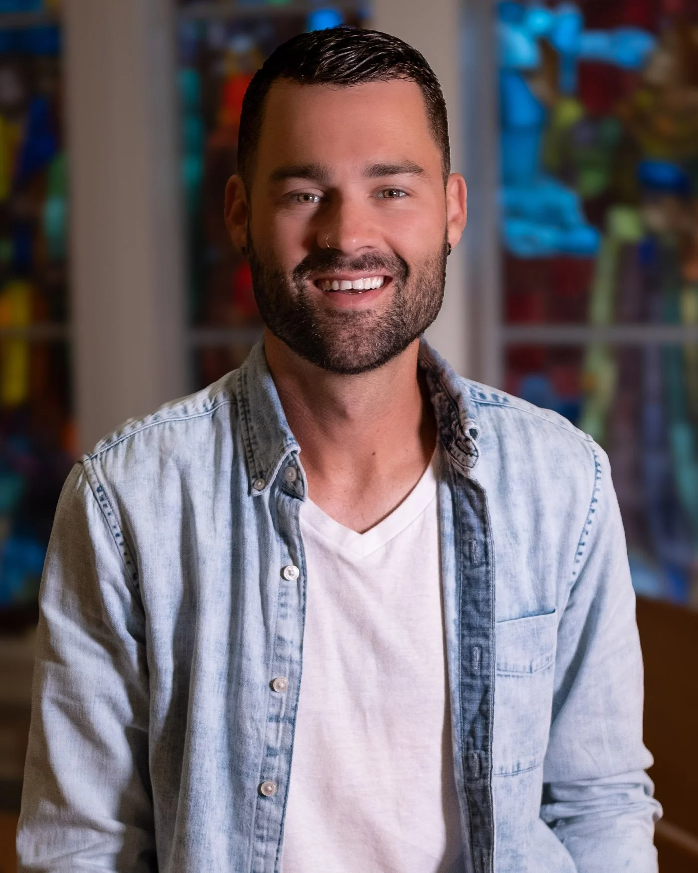 A man with dark hair and a beard, smiling, wearing a light denim shirt over a white t-shirt, standing indoors in front of a blurred background with colorful objects.