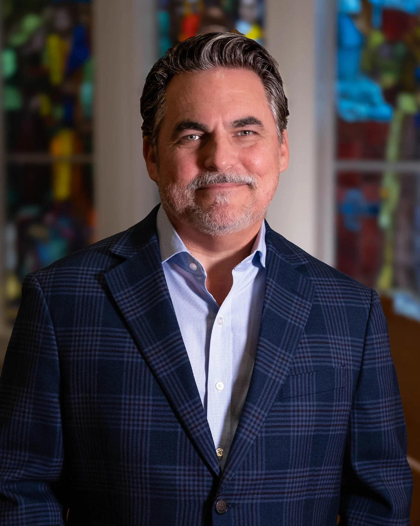 A middle-aged man with gray hair and a beard dressed in a plaid suit jacket and a light blue collared shirt standing indoors with a blurred colorful background.