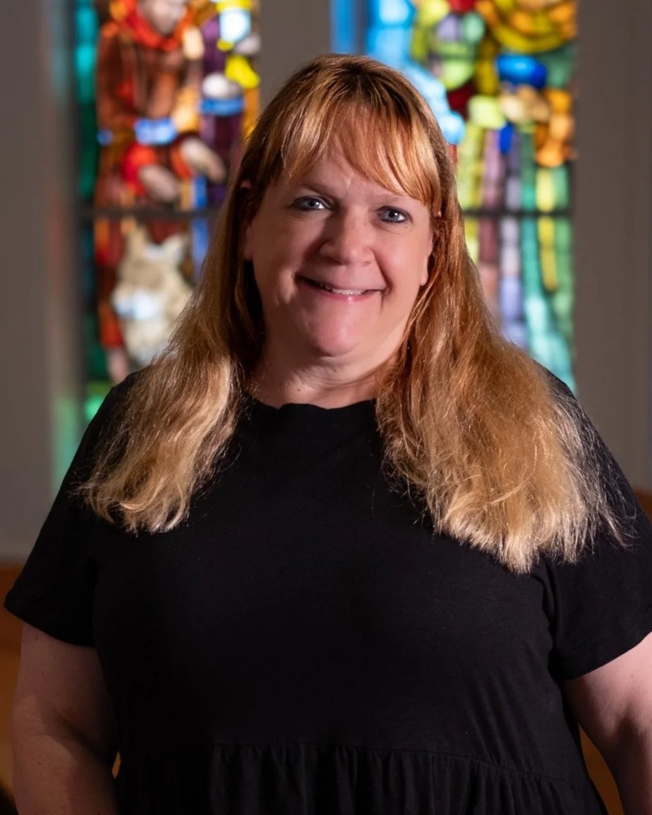 A woman with reddish hair and a blonde ponytail, smiling, wearing a navy blue floral top, standing indoors with large windows in the background.