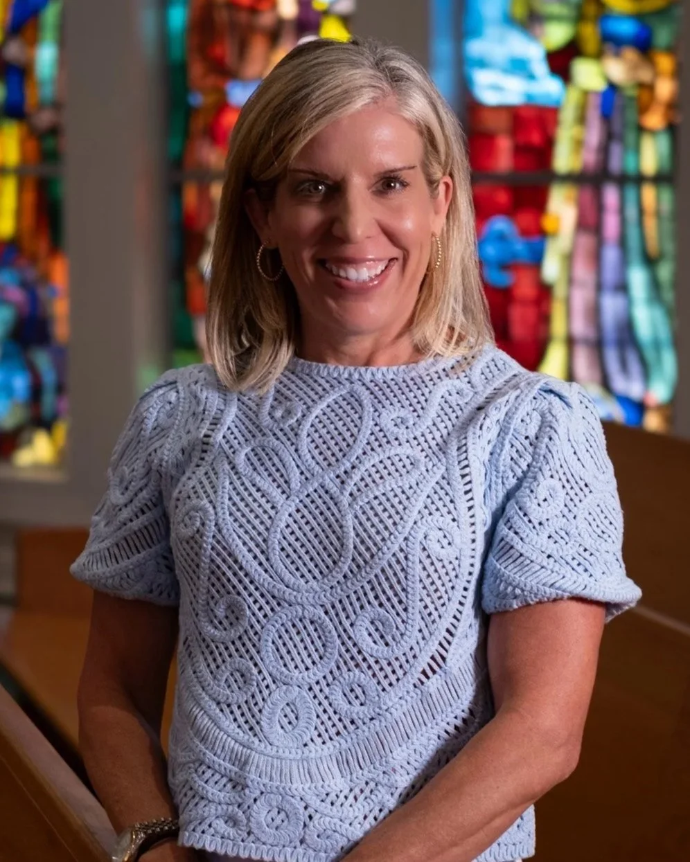 A smiling woman with shoulder-length blonde hair wearing a beige t-shirt and gold hoop earrings, standing inside a building with large windows and a door in the background.