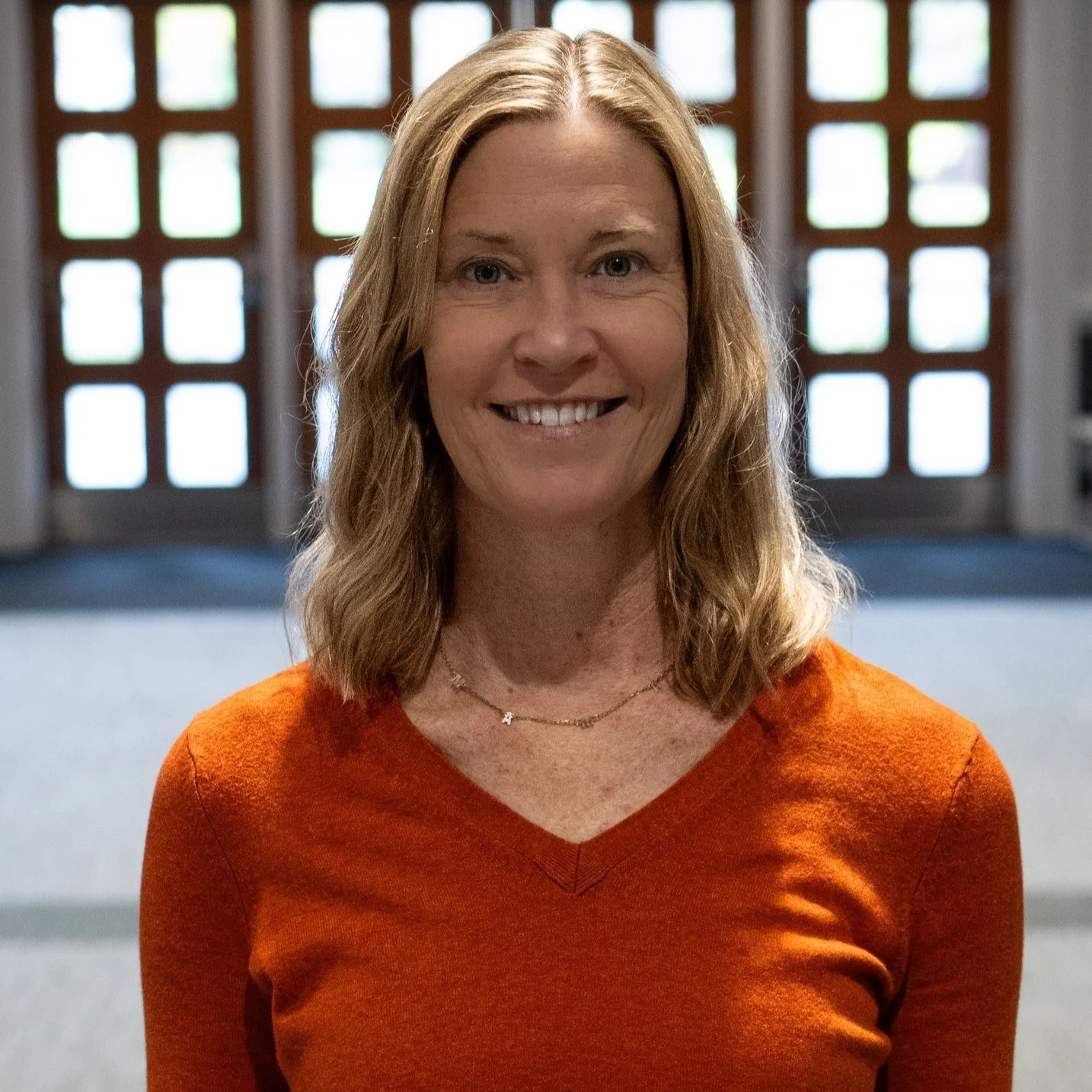 A woman with shoulder-length blonde hair in an orange top smiling, standing indoors with a backdrop of large grid-style windows.