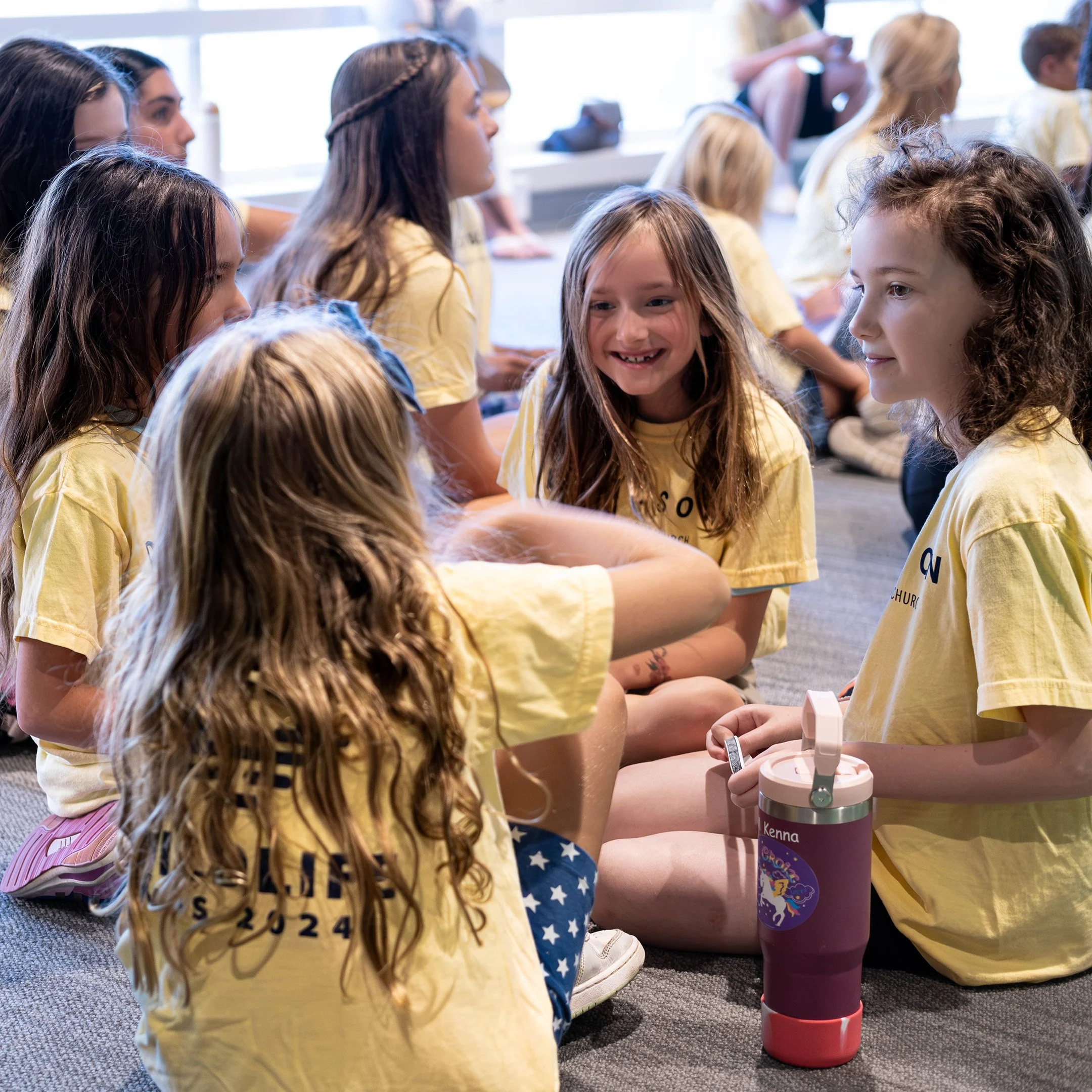Group of children sitting on the floor, wearing yellow t-shirts, engaged in conversation and smiling during a group activity.