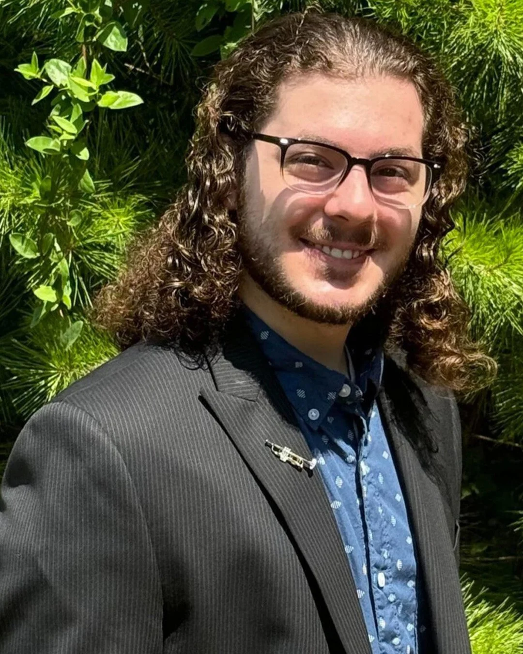 A man with long curly hair, beard, glasses, and wearing a suit with a blue patterned shirt, smiling outdoors with green foliage in the background.