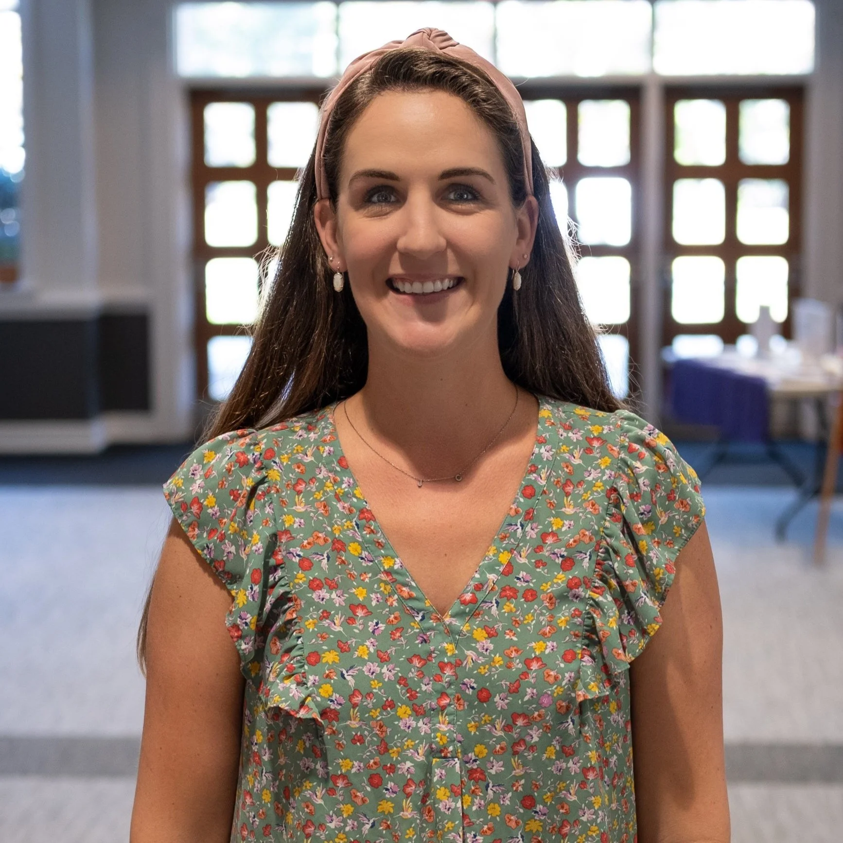 A woman with long brown hair, wearing a pink headband and a floral dress, smiling in a brightly lit room with large windows and wooden frames.