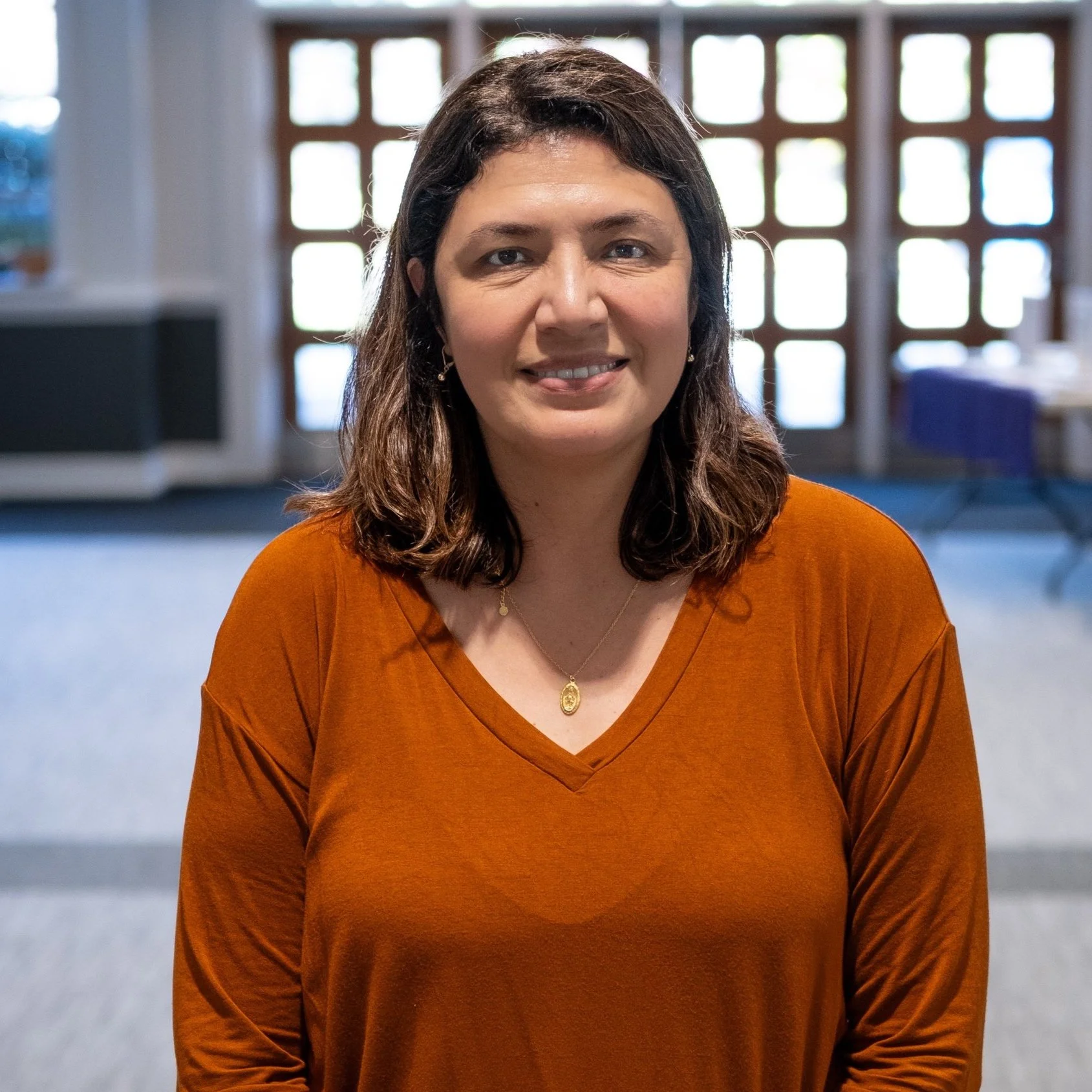 A woman with shoulder-length brown hair smiling, wearing an orange V-neck shirt and a gold necklace, in a room with large windows and a table in the background.