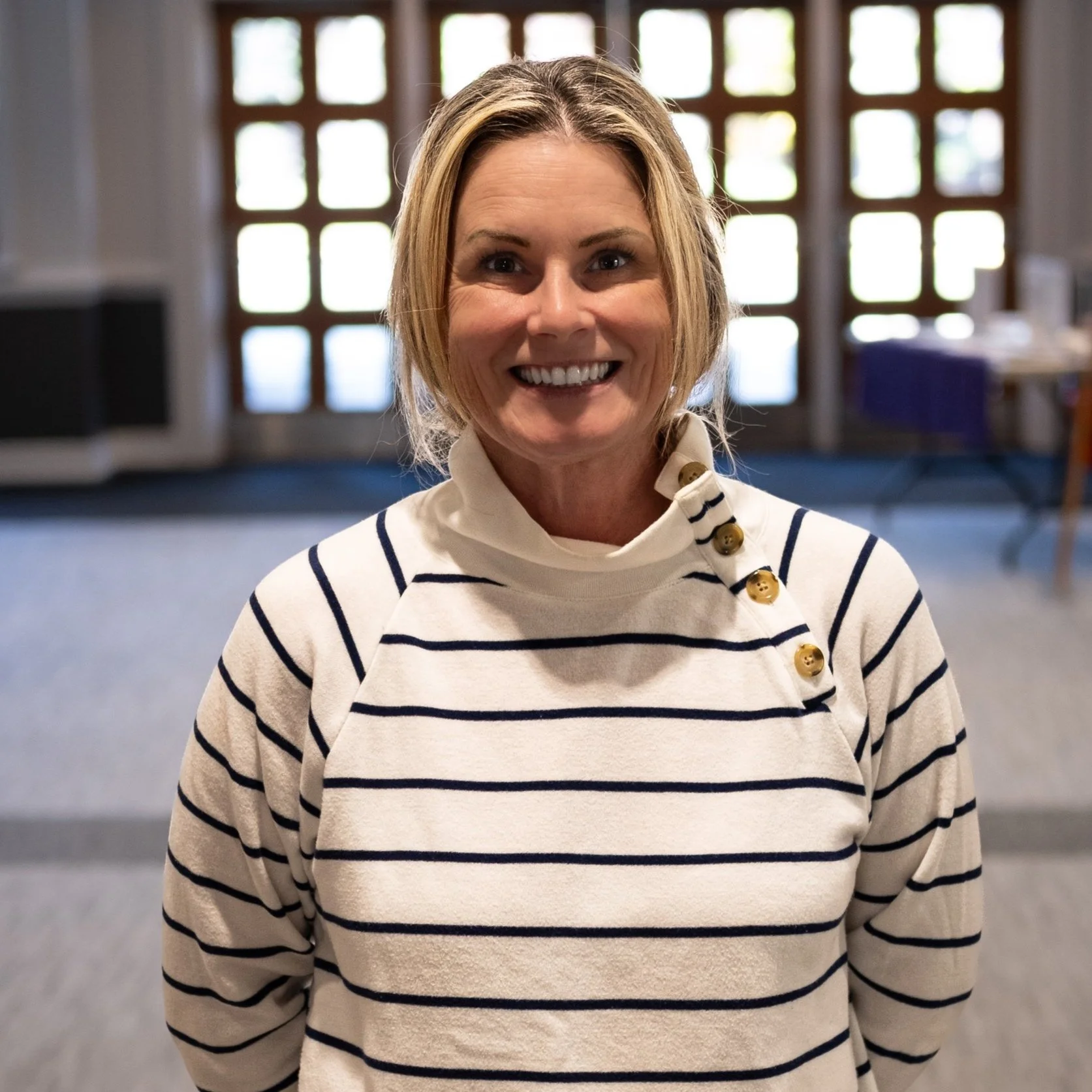 A smiling woman with shoulder-length blonde hair wearing a cream-colored sweater with navy horizontal stripes and decorative buttons on the shoulder, standing indoors with a background of windows and blurred interior elements.