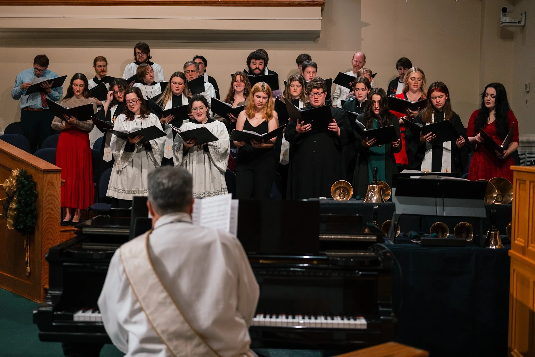 A choir performing on stage with a conductor, with a pianist at a grand piano in the foreground, during a holiday or Christmas event.