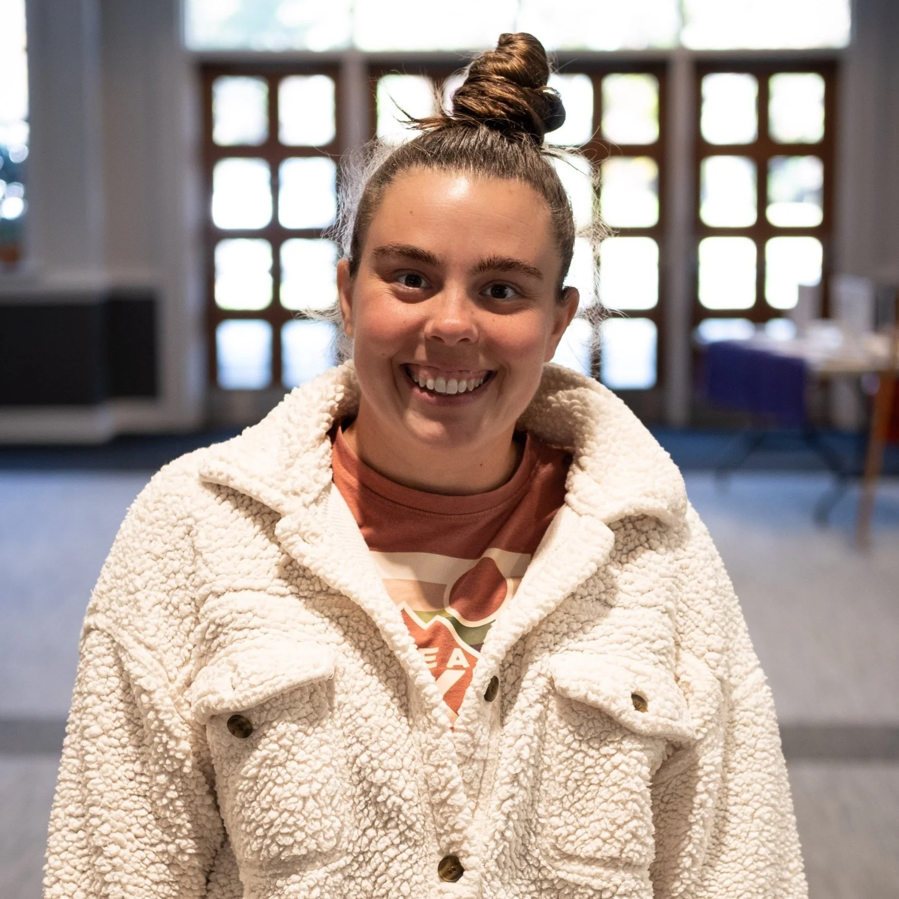 Young woman with a top knot hairstyle, smiling, wearing a beige textured fleece jacket inside a room with large windows.