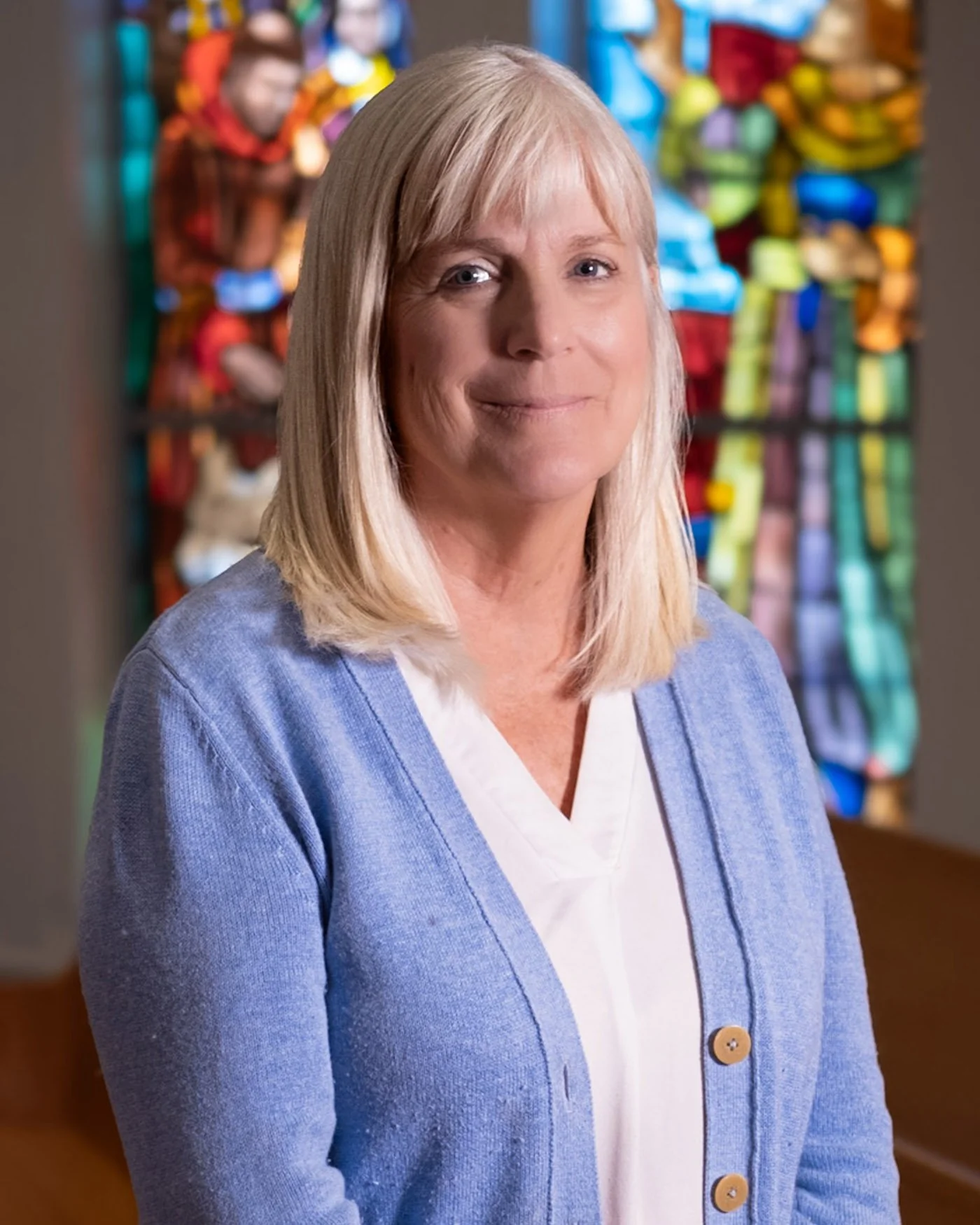 A middle-aged woman with blonde hair, wearing a blue cardigan over a white shirt, standing indoors with a stained glass window in the background.