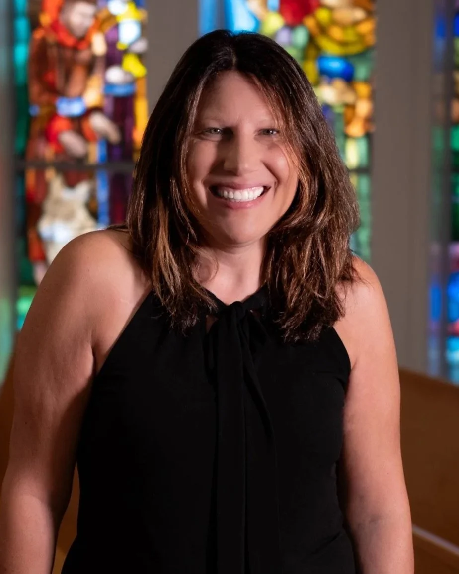 A woman with shoulder-length brown hair smiling at the camera, wearing a black cardigan and a red and white patterned top, inside a room with a large window with grid-like panes in the background.