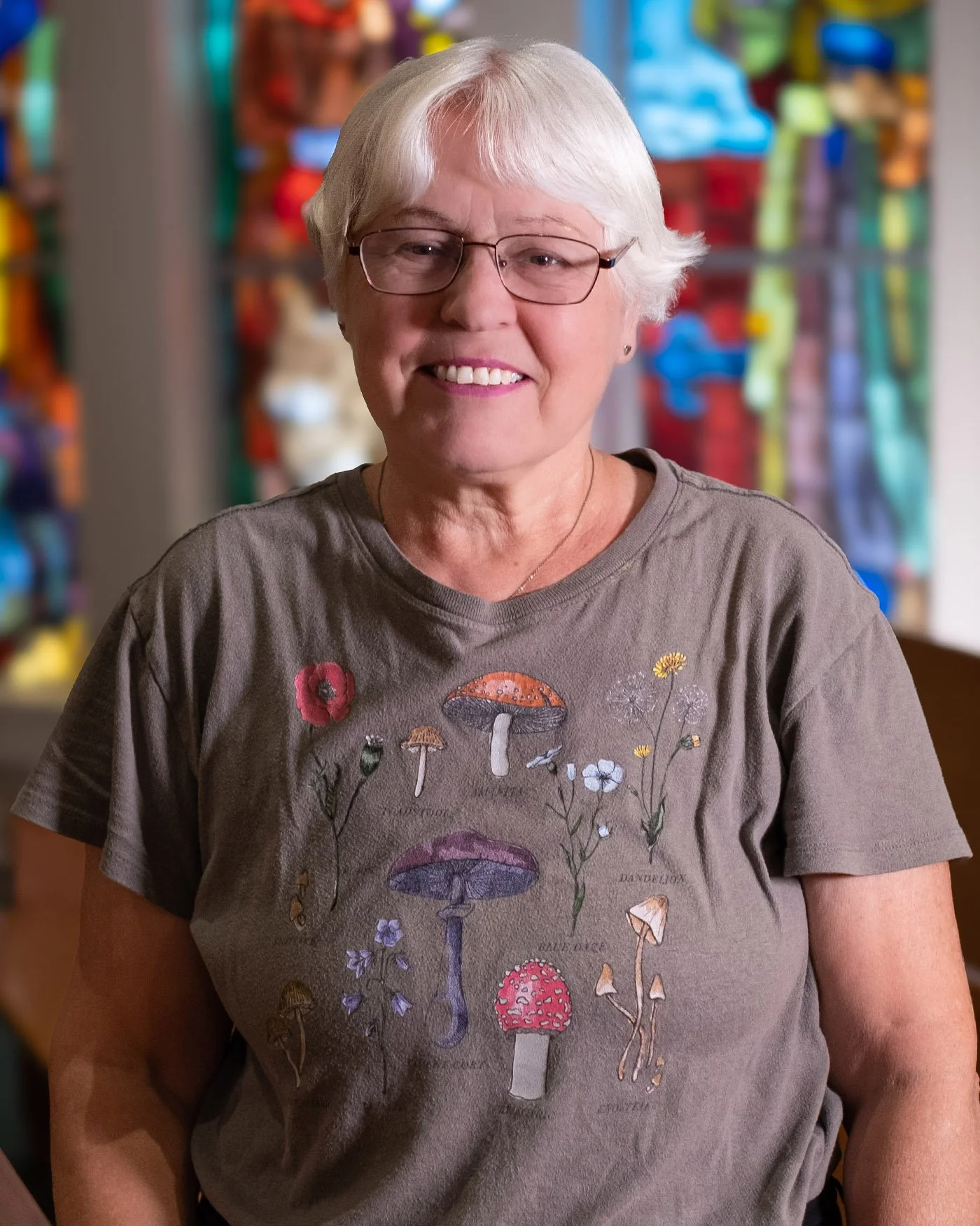 A smiling elderly woman with short white hair and glasses wearing a gray T-shirt with colorful mushroom and flower illustrations, standing indoors with a multicolored, artful background.
