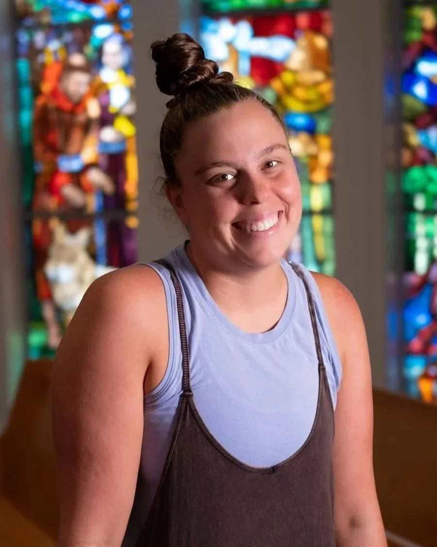 A young woman with brown hair in a top bun, smiling, wearing a cream fleece jacket, standing indoors with large grid windows in the background.