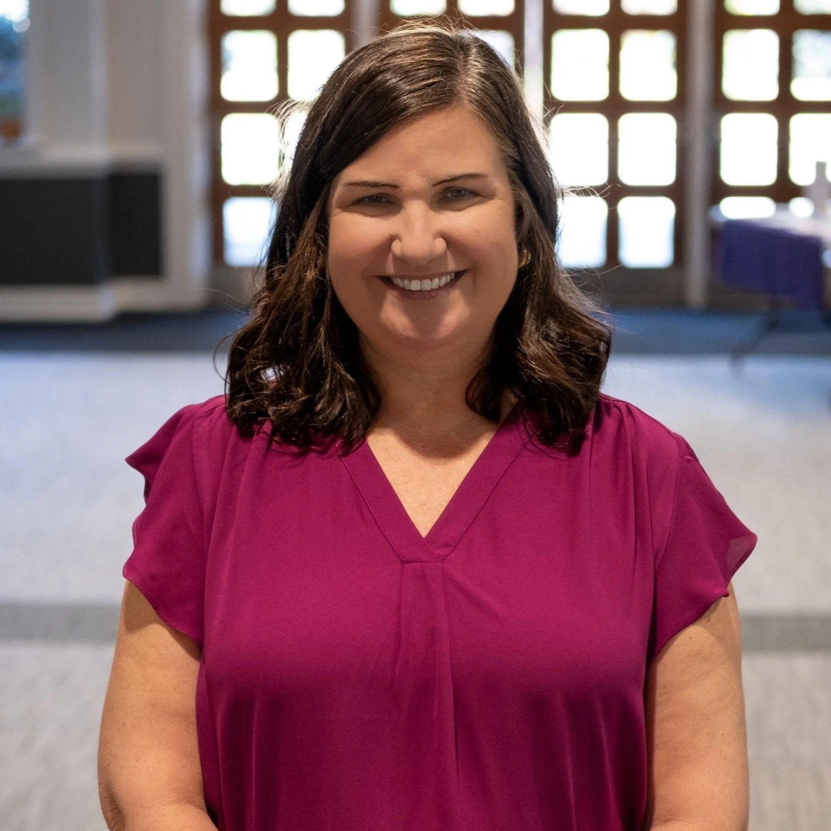 A smiling woman with shoulder-length dark hair, wearing a magenta blouse, standing indoors in front of large windows with lattice-patterned frames.