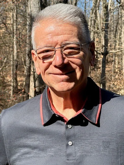 An older man with gray hair and glasses, smiling outdoors in a wooded area, wearing a dark collared shirt.