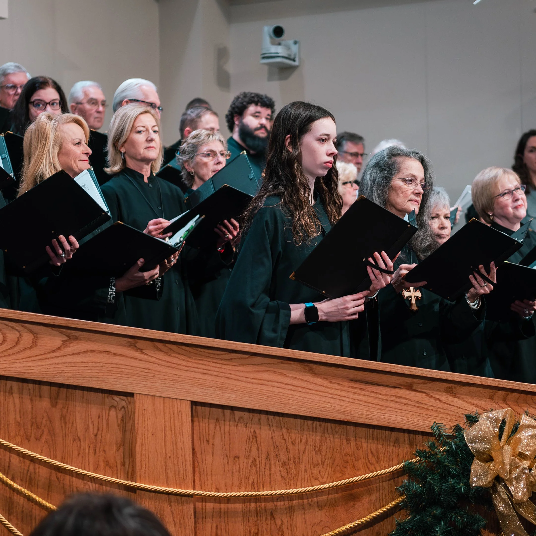A group of choir singers, mostly women, standing on a stage, holding sheet music, dressed in black robes with a Christmas decoration in the foreground.