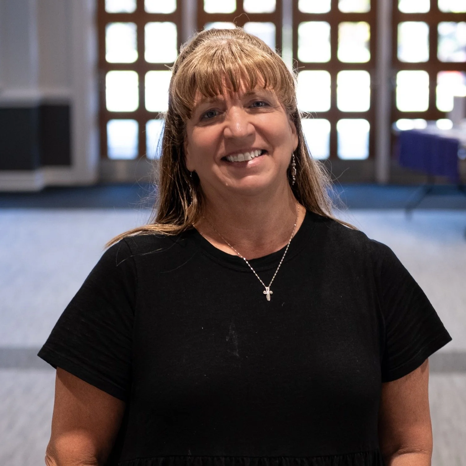 A woman with light brown hair, wearing a black shirt, smiling inside a room with a patterned window in the background.