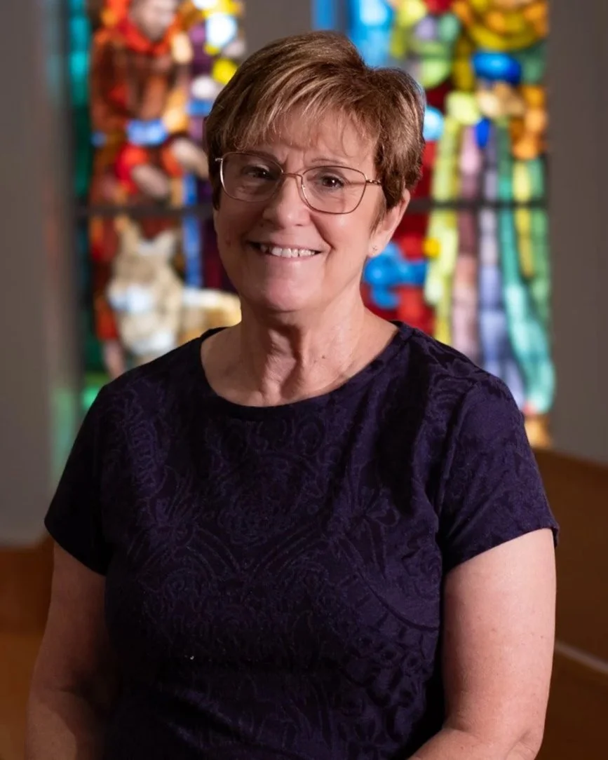 An older woman with short light brown hair, glasses, and a pink shirt with eyelet details on the shoulders, smiling indoors in front of large windows.