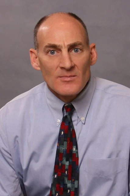 A man with a serious expression, wearing a light blue dress shirt and a patterned tie, against a plain gray background.
