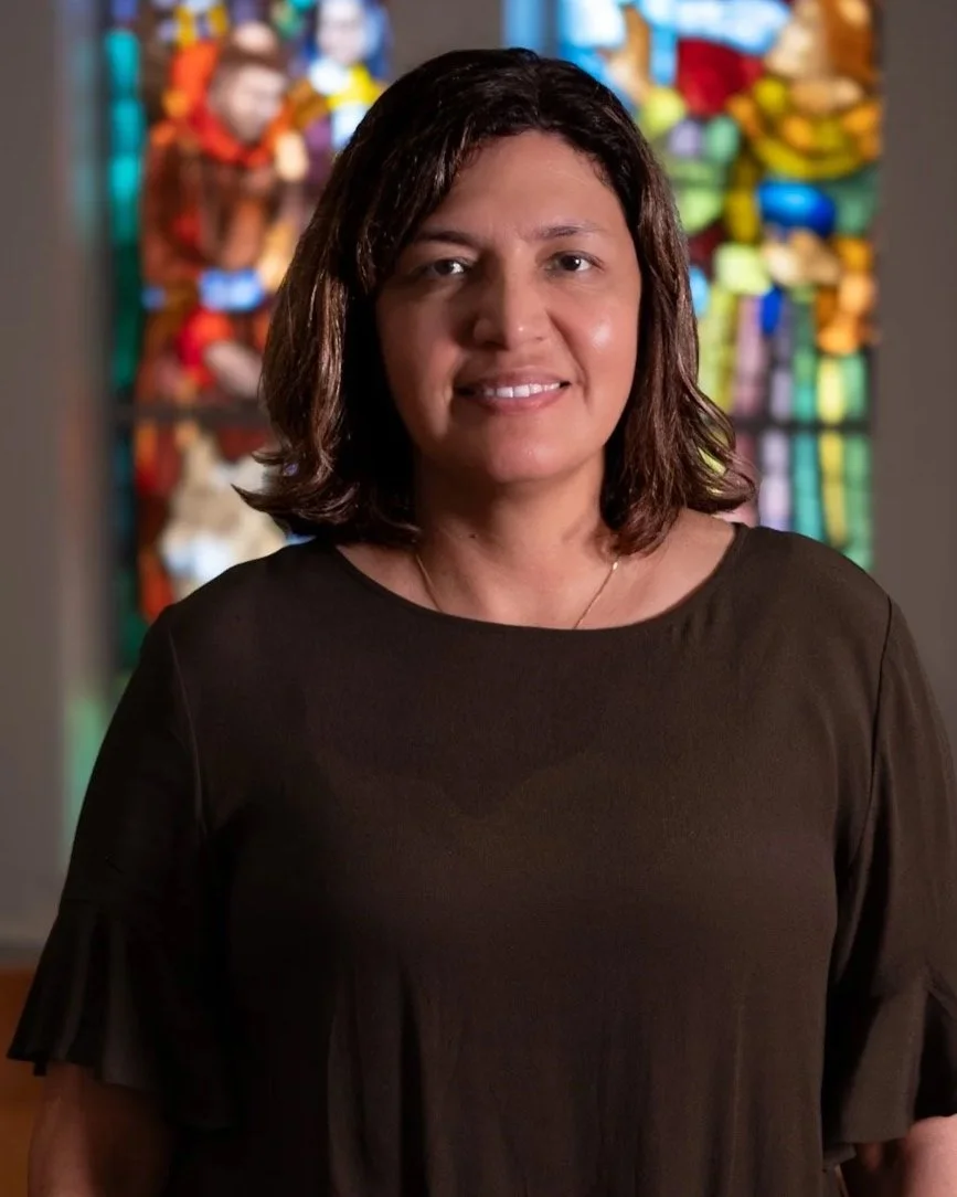 A woman with shoulder-length brown hair smiling, wearing an orange V-neck shirt and a gold necklace, in a room with large windows and a table in the background.