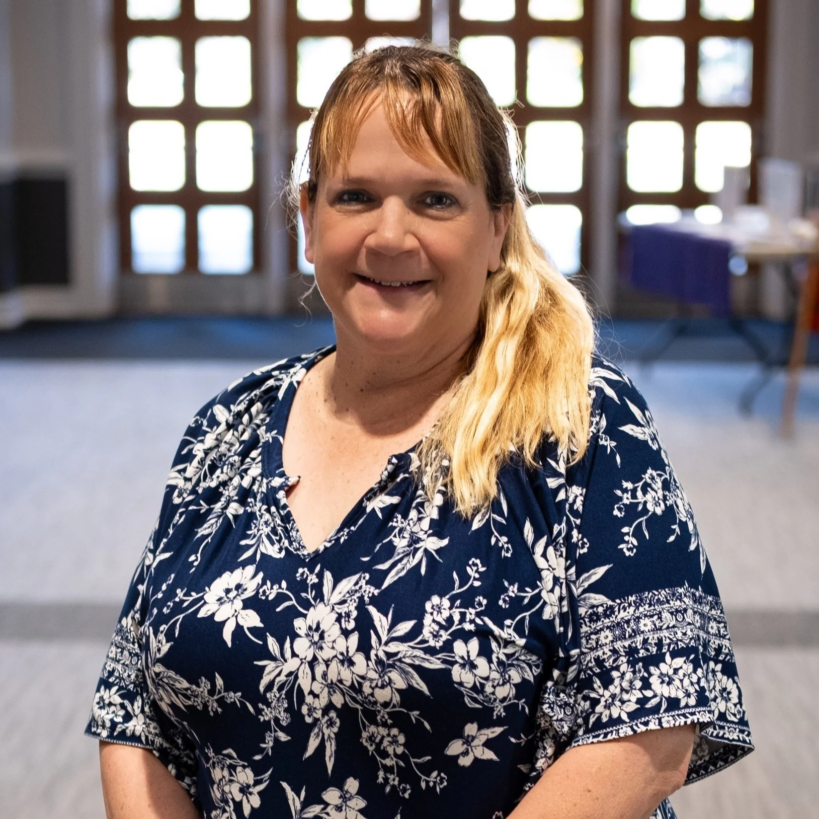 A woman with reddish hair and a blonde ponytail, smiling, wearing a navy blue floral top, standing indoors with large windows in the background.