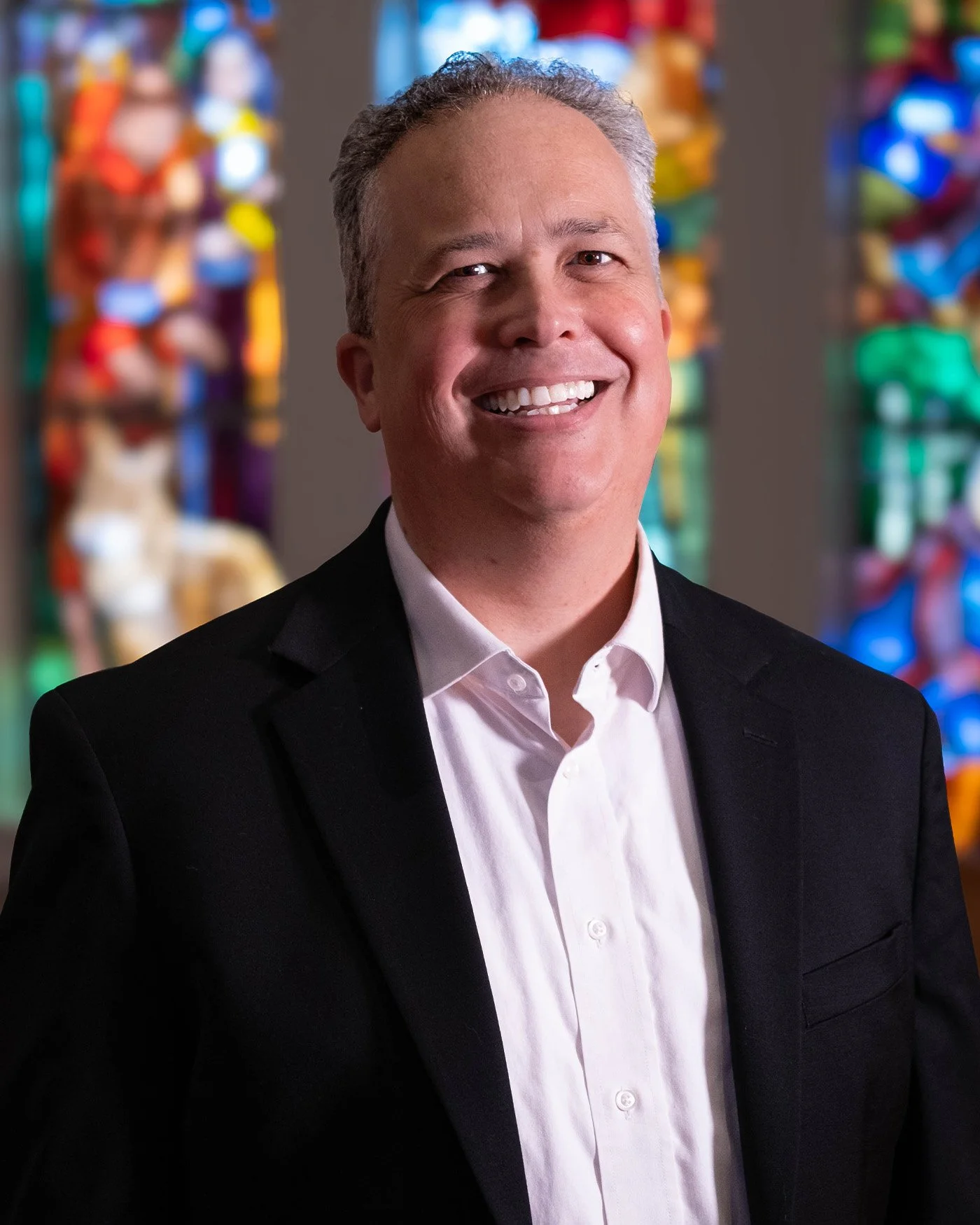 A smiling man in a black suit and white shirt standing in front of colorful stained glass windows.