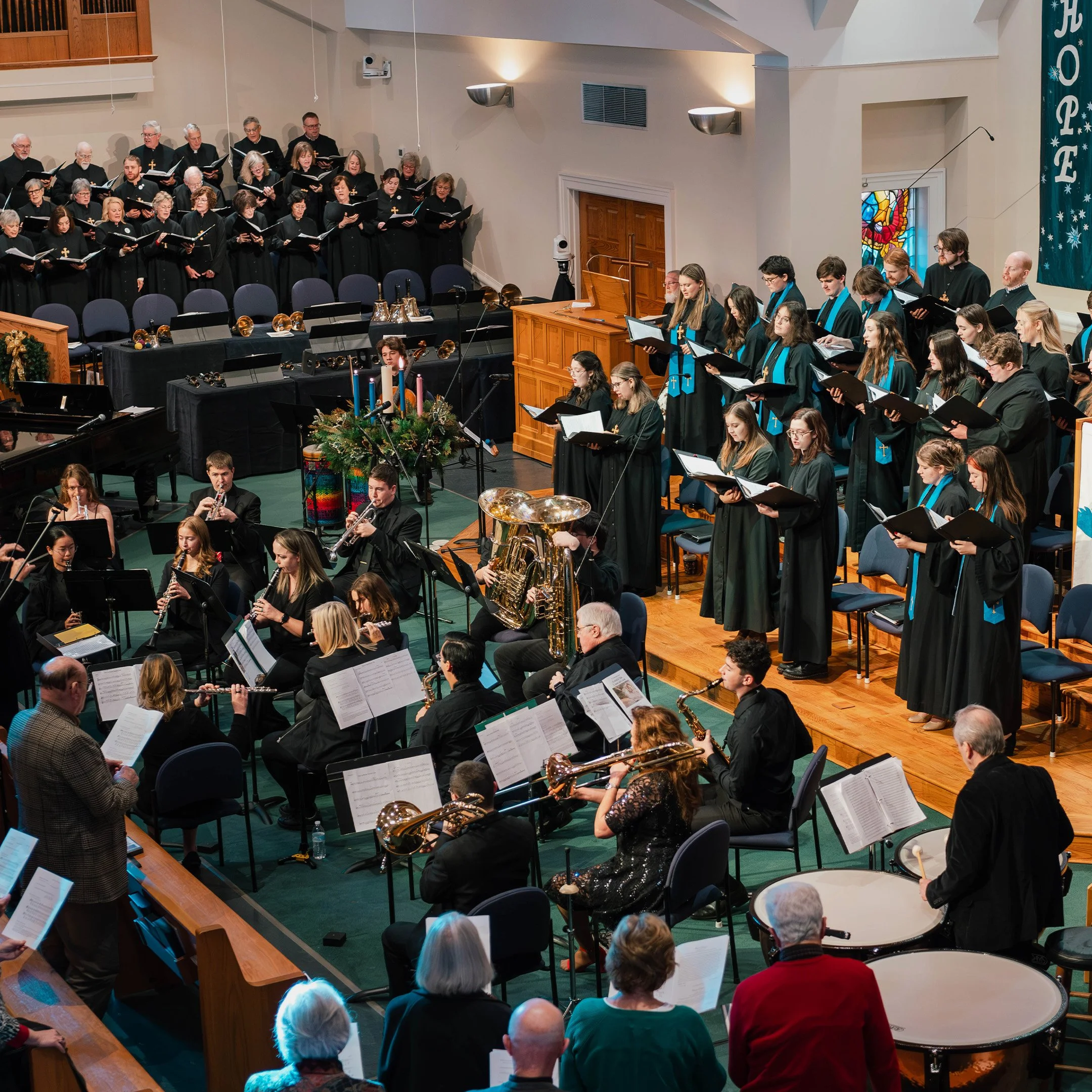 A large choir and orchestra performing inside a church with stained glass windows. The choir is positioned on risers at the back, dressed in black, with some wearing blue stoles. The orchestra, seated in front, includes various instruments like flute