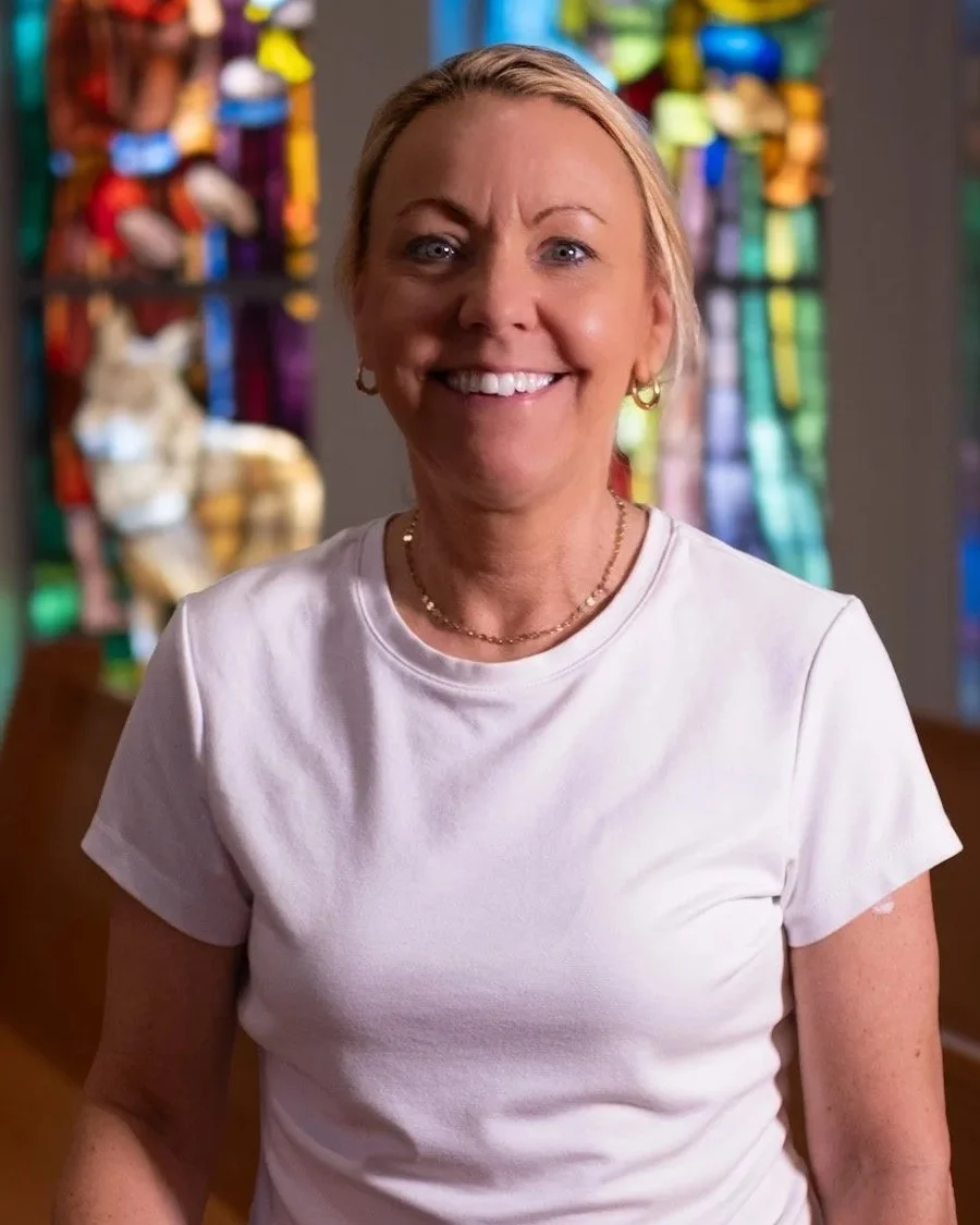 Smiling woman with light brown hair in a ponytail, wearing a white button-up shirt and a pink quilted vest, standing inside a building with large window panes in the background.