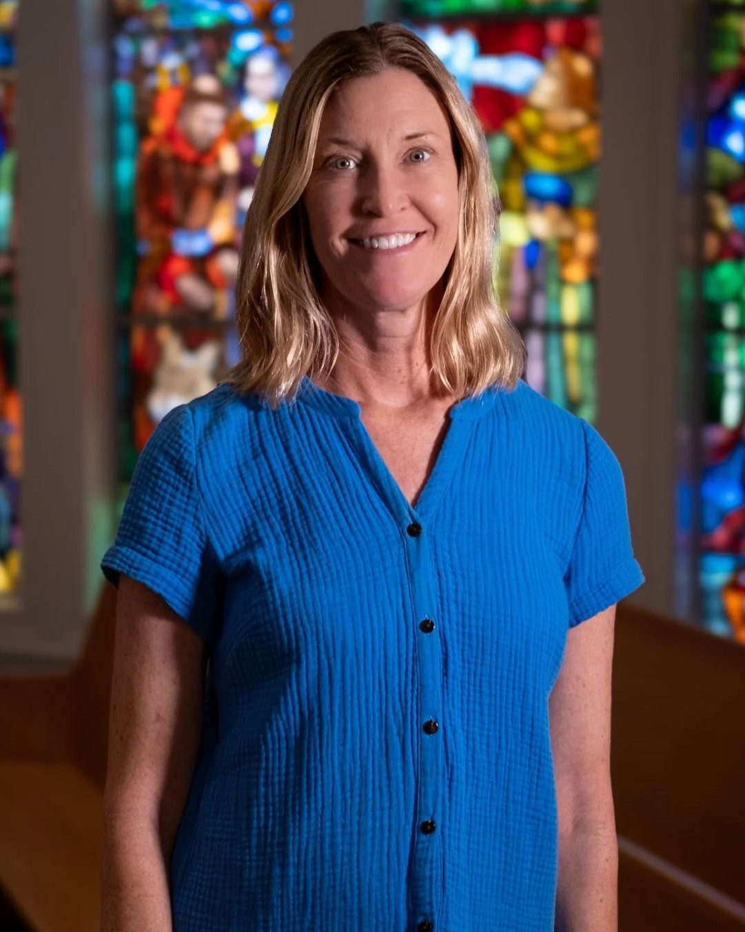 A woman with shoulder-length blonde hair in an orange top smiling, standing indoors with a backdrop of large grid-style windows.
