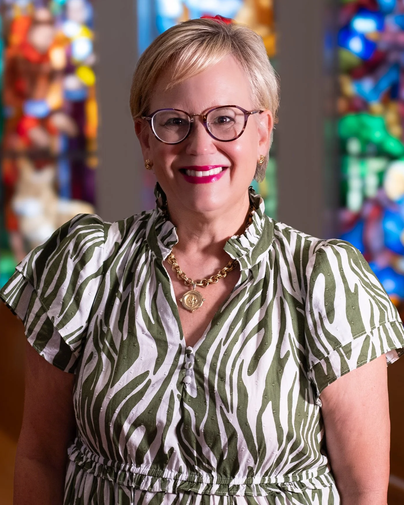 A woman with short blonde hair and glasses smiling in front of a colorful background.