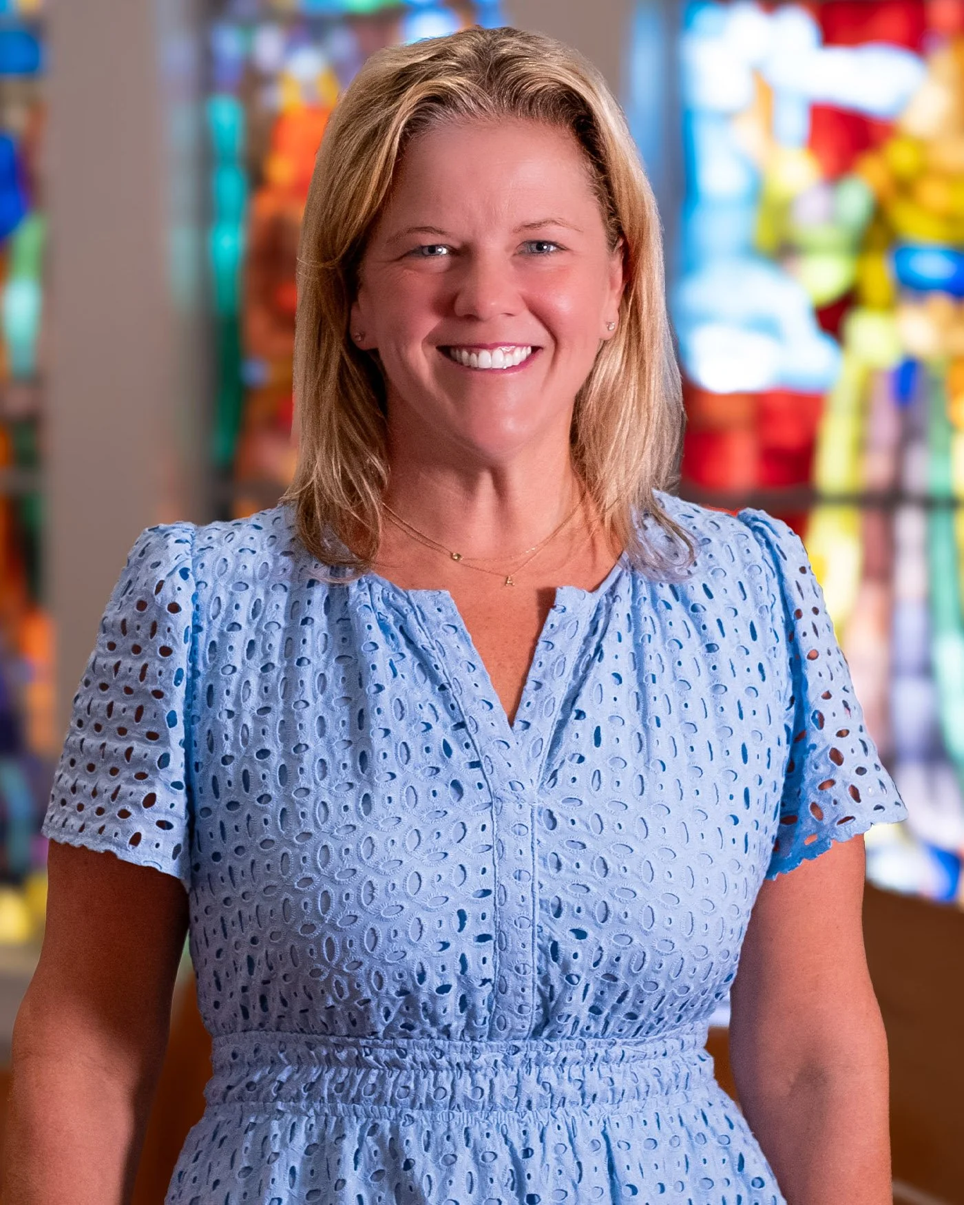 A smiling woman with blonde hair wearing a light blue, eyelet dress stands in front of a colorful store display.