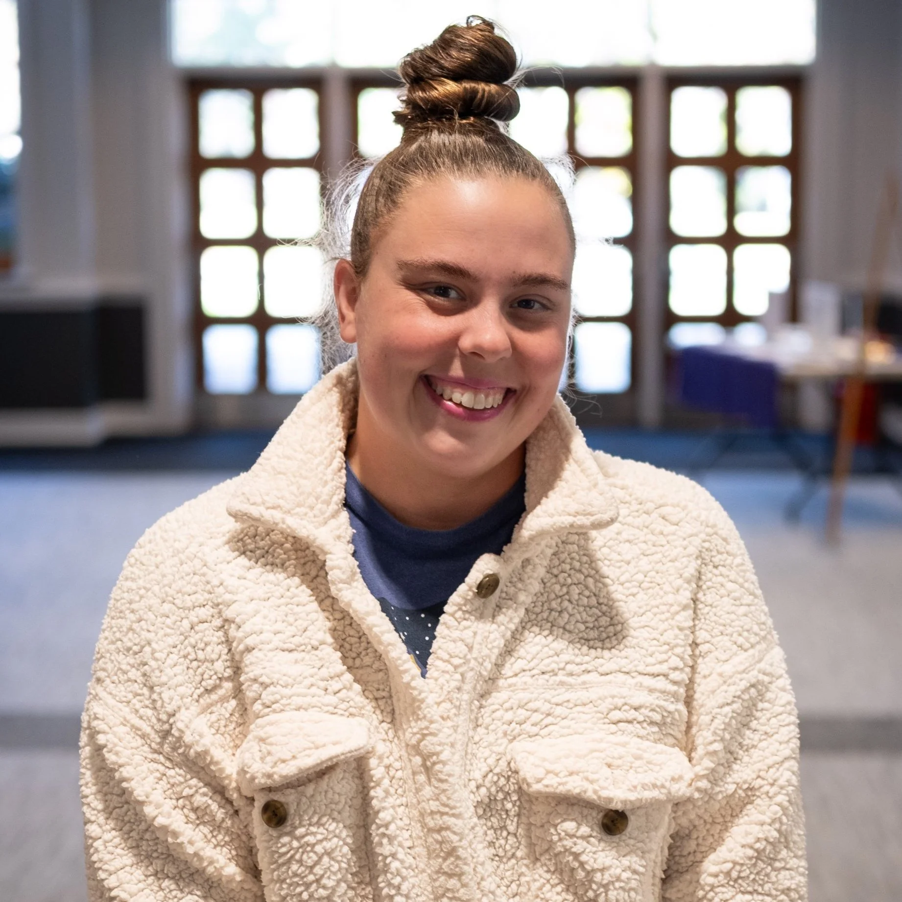 A young woman with brown hair in a top bun, smiling, wearing a cream fleece jacket, standing indoors with large grid windows in the background.