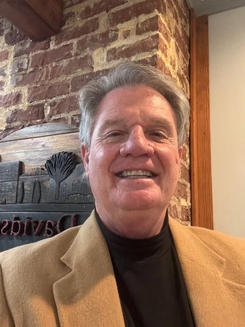 A smiling man with gray hair wearing a tan blazer and a black turtleneck, indoor setting with brick wall and wooden accents in the background.